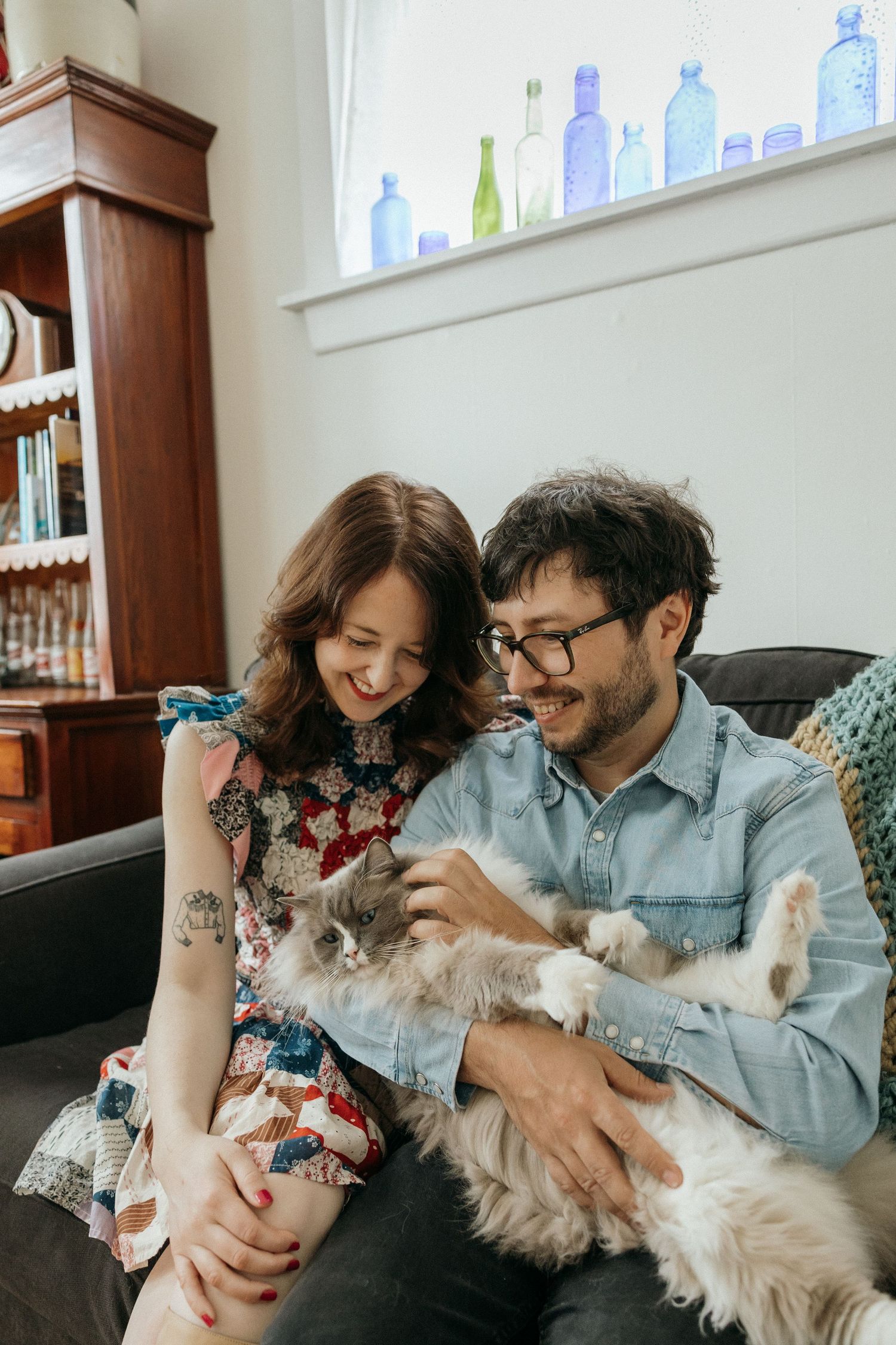 A couple plays with their cats while sitting on a couch in a bright living room.