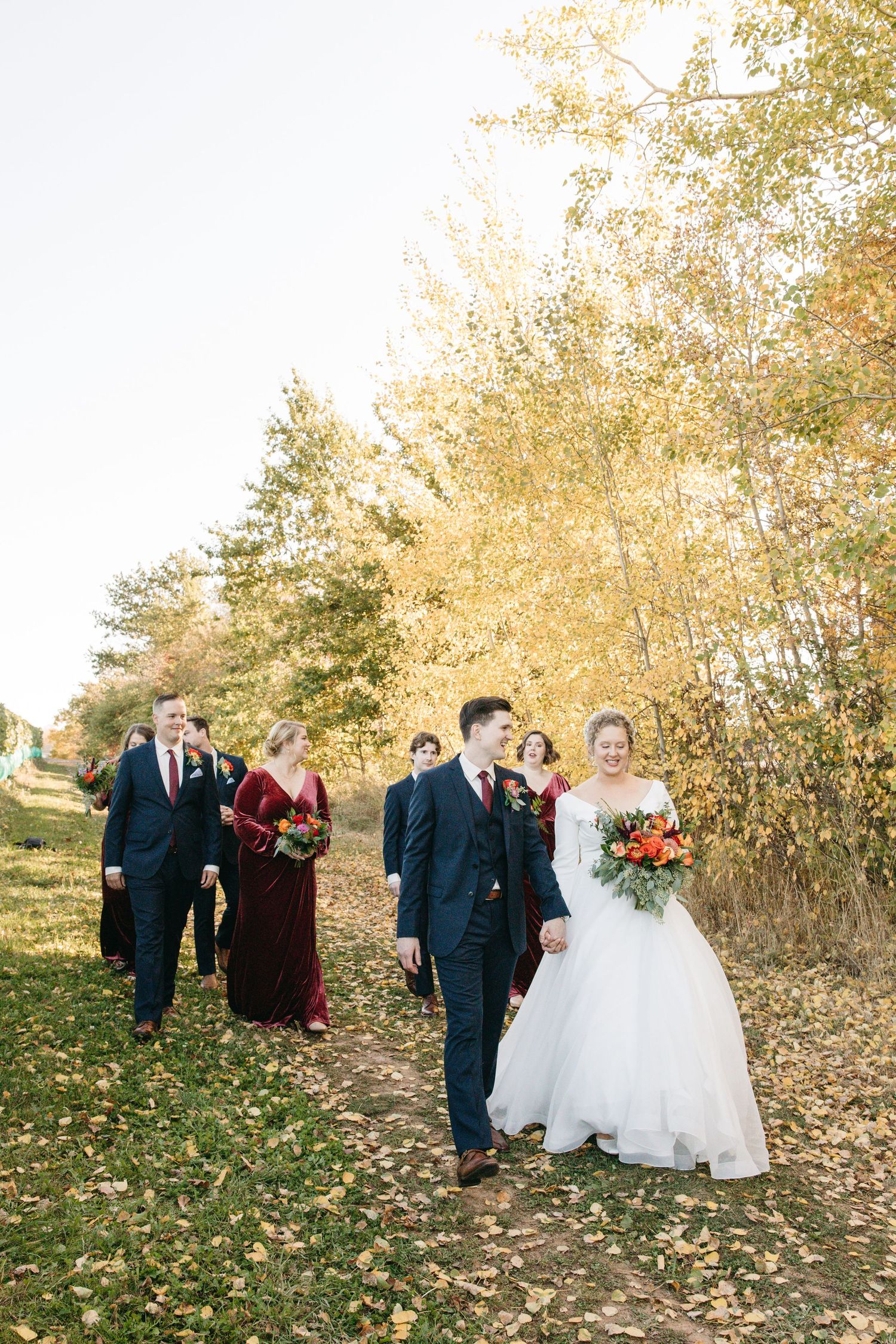 Wedding party walks along a fall path lined with golden trees and scattered leaves.