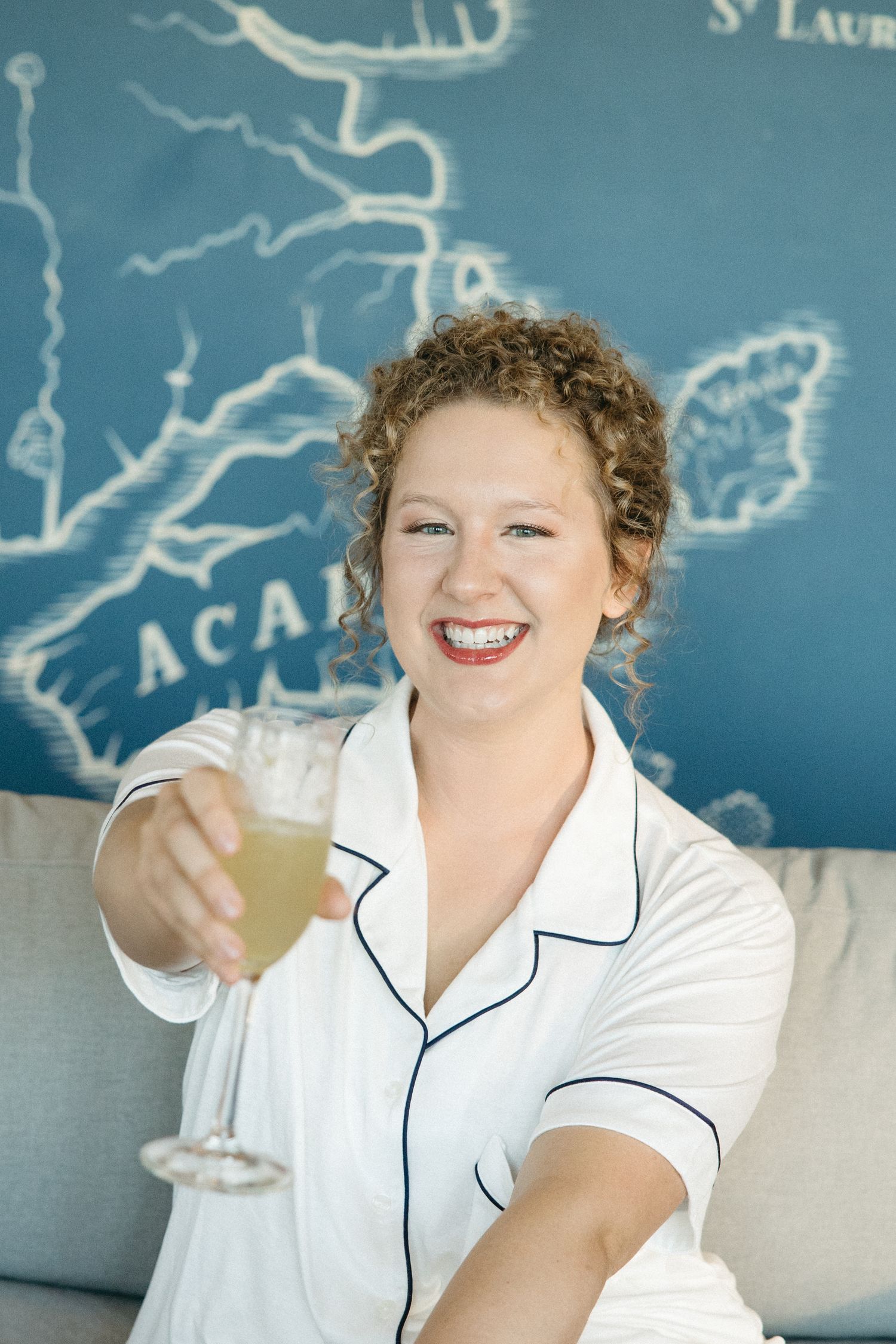 Person in white pajamas with navy piping smiles while holding a drink against blue wall art.
