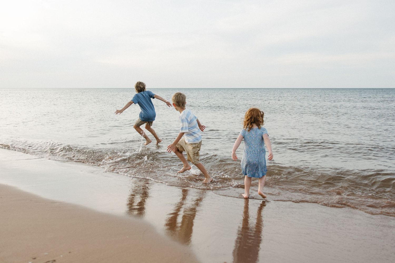 Three children playing in ocean waves on a sandy beach on a cloudy day.