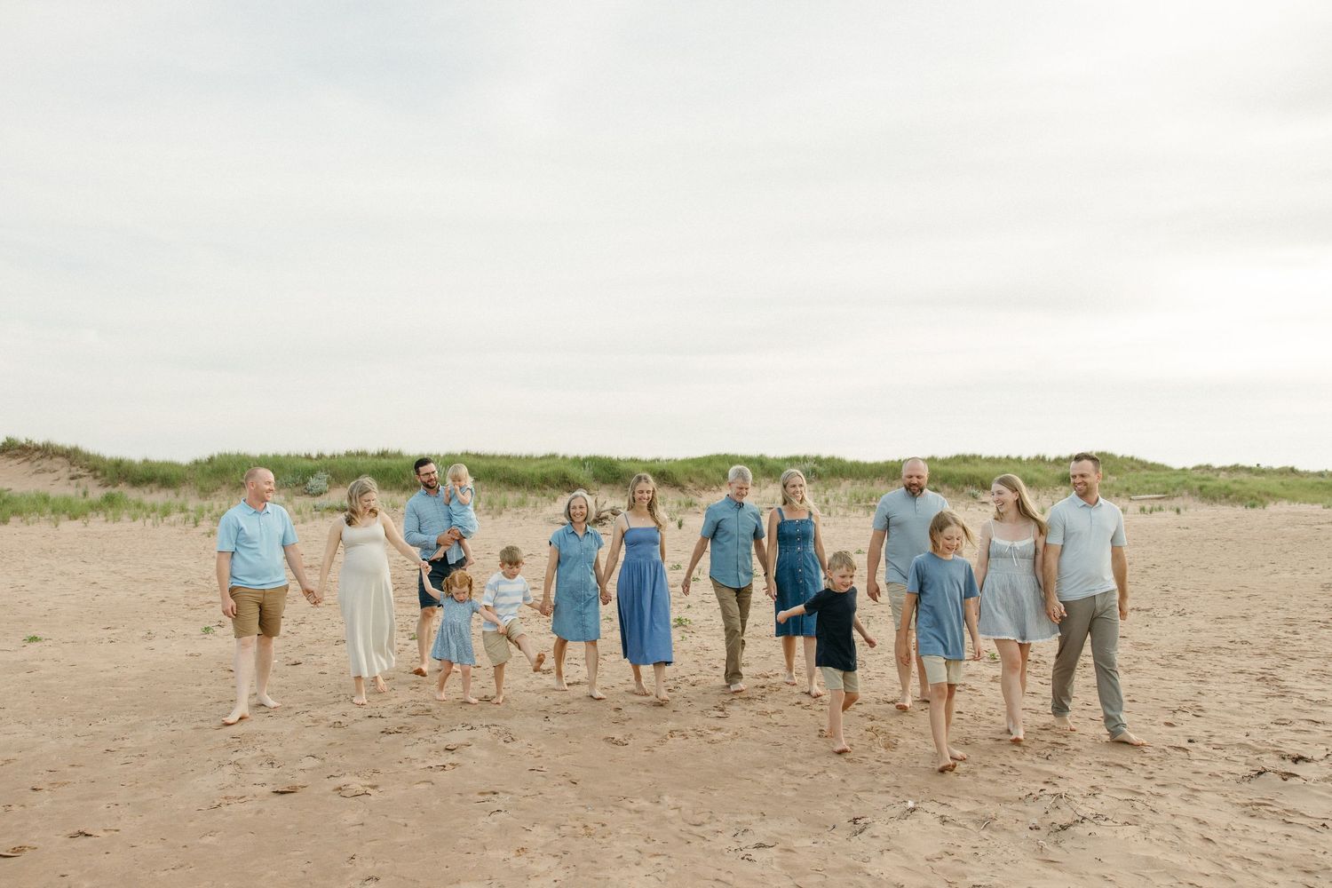 Large extended family holds hands in a line on sandy beach with grass dunes during an outdoor portrait.