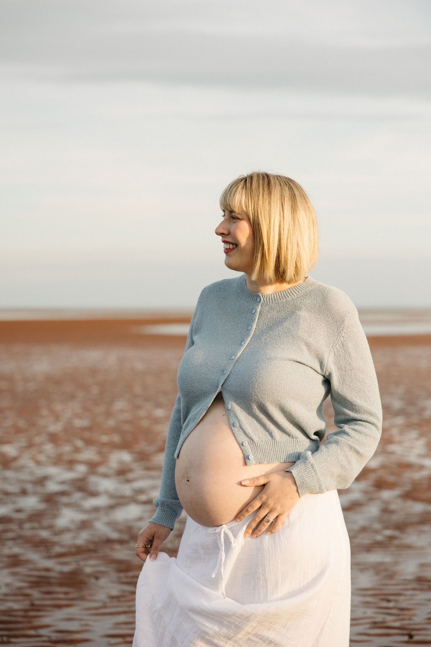 Glowing pregnant blonde woman in blue cardigan holds her bare bump while standing on a sandy beach.
