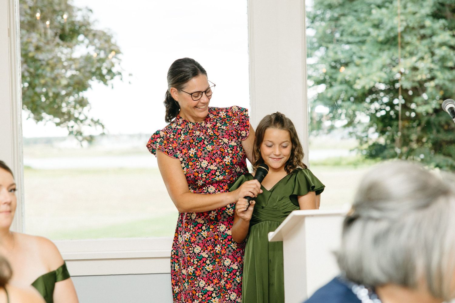 Woman in floral dress helps a young girl in green dress read during a wedding ceremony.