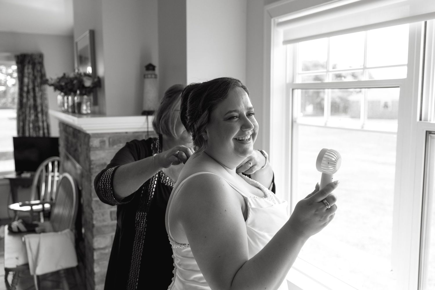 Bride smiling as bridesmaid helps with dress in black and white wedding preparation photo indoors.