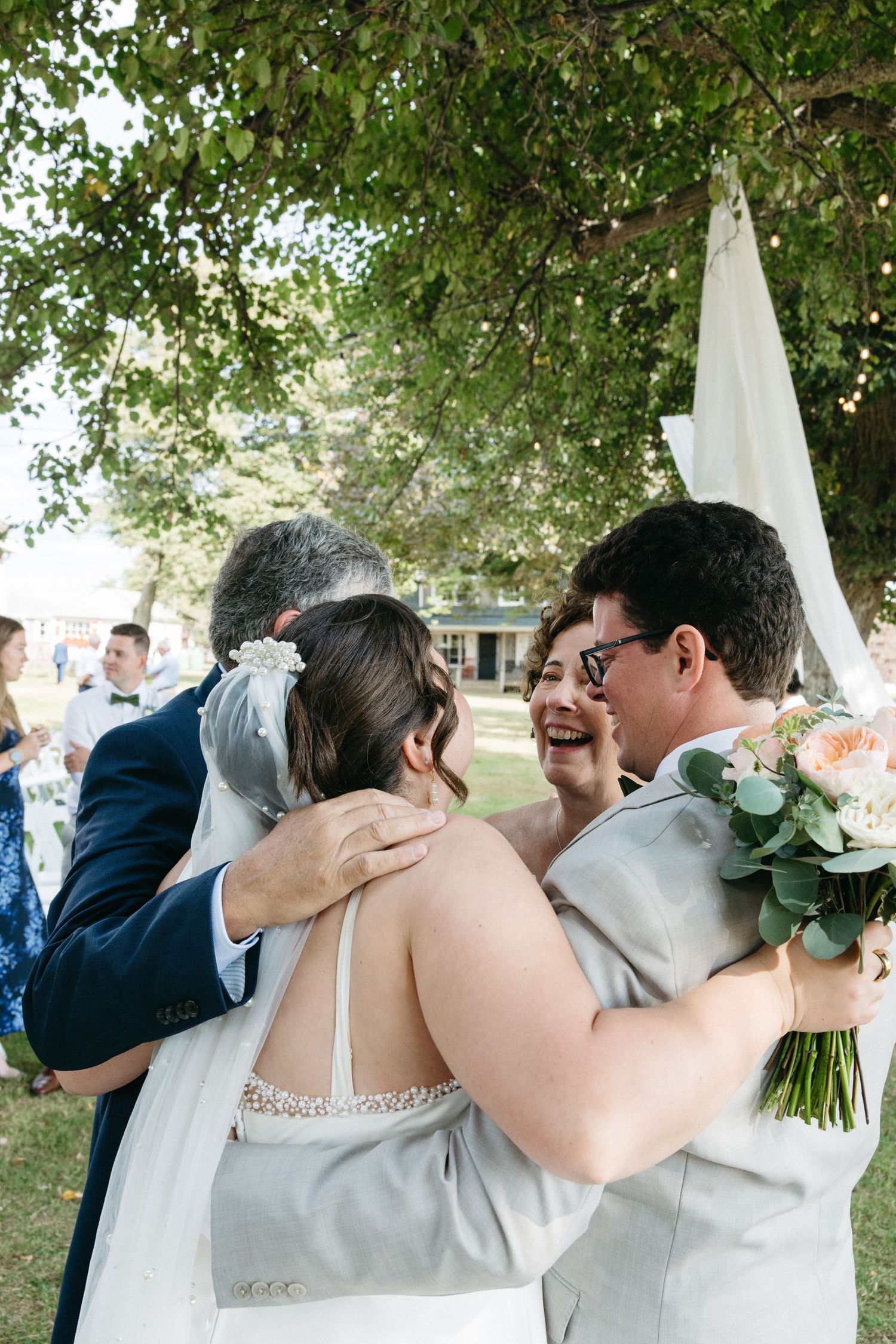 Bride and groom joyfully embracing family members under a large tree at an outdoor wedding ceremony.