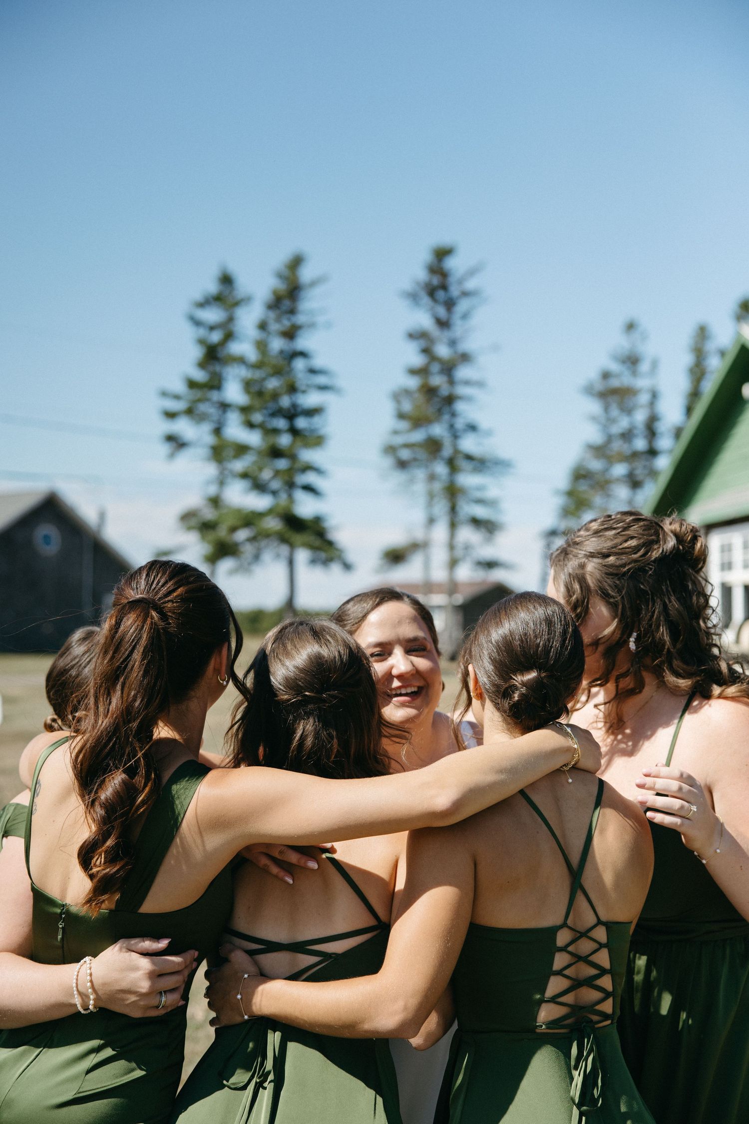 Bridesmaids in green dresses share a group hug outdoors on a sunny wedding day.