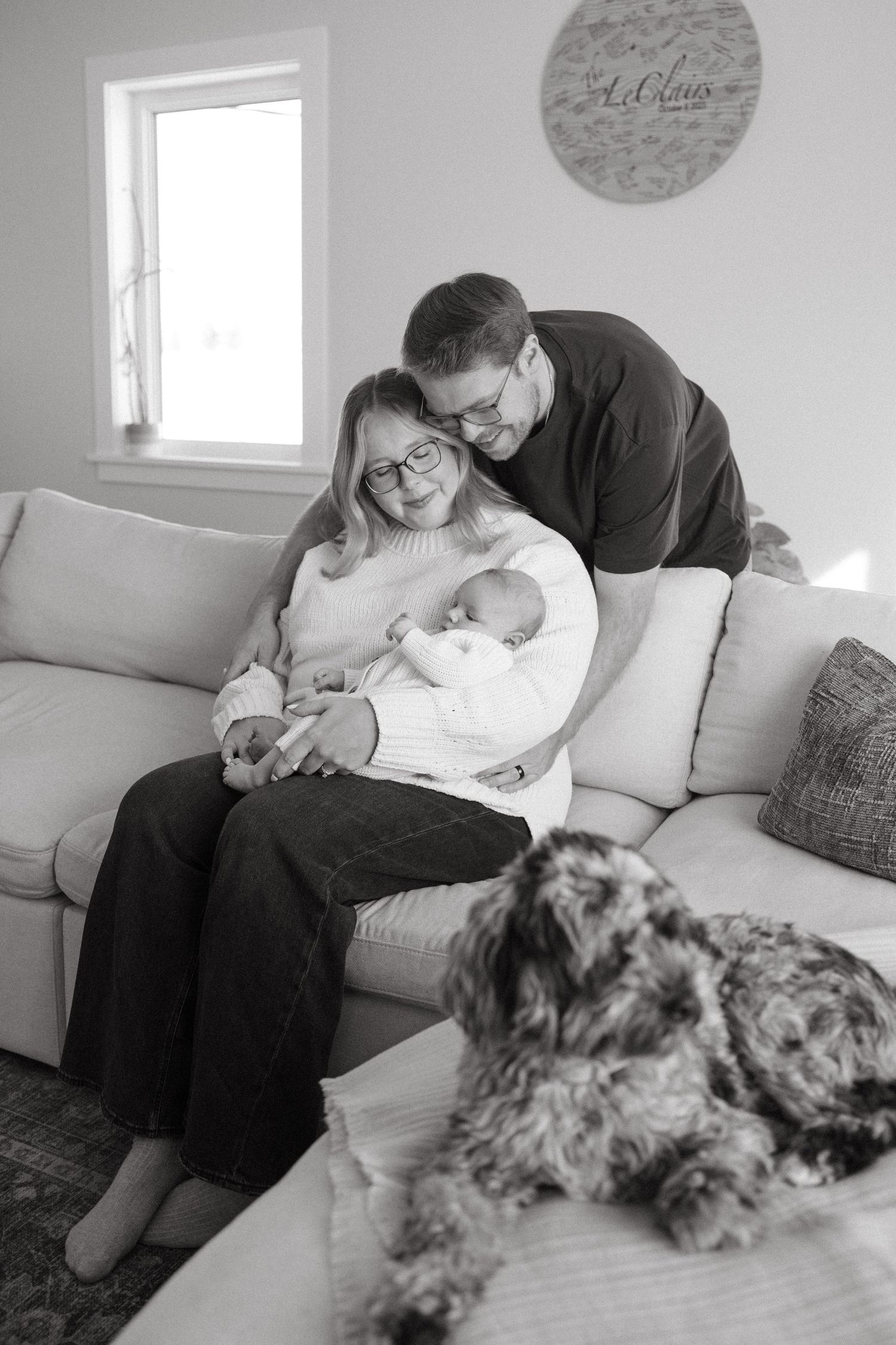 A family shares an intimate moment on their gray couch with their baby and dog in a bright living room.