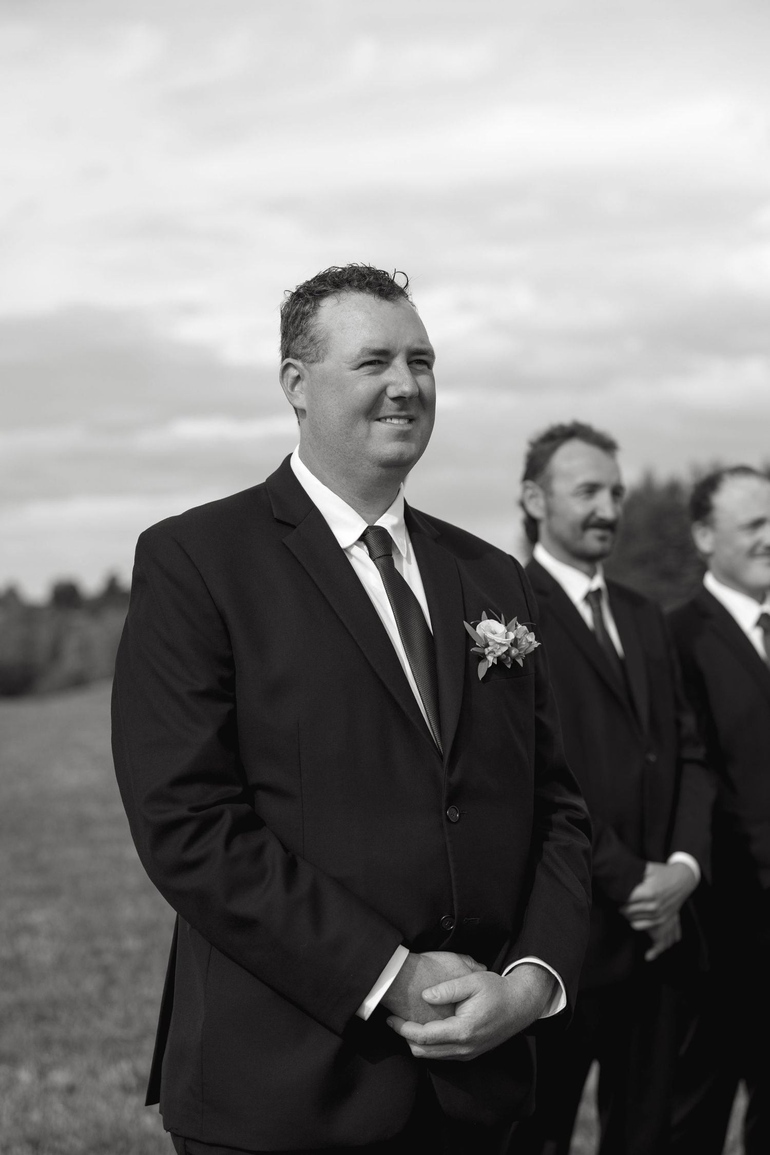 Groomsmen in black suits sharing a laugh in an outdoor setting.