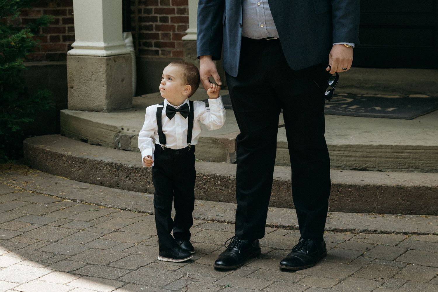 Close-up view of formal dress shoes and pant legs standing on brick pavement at an outdoor event.