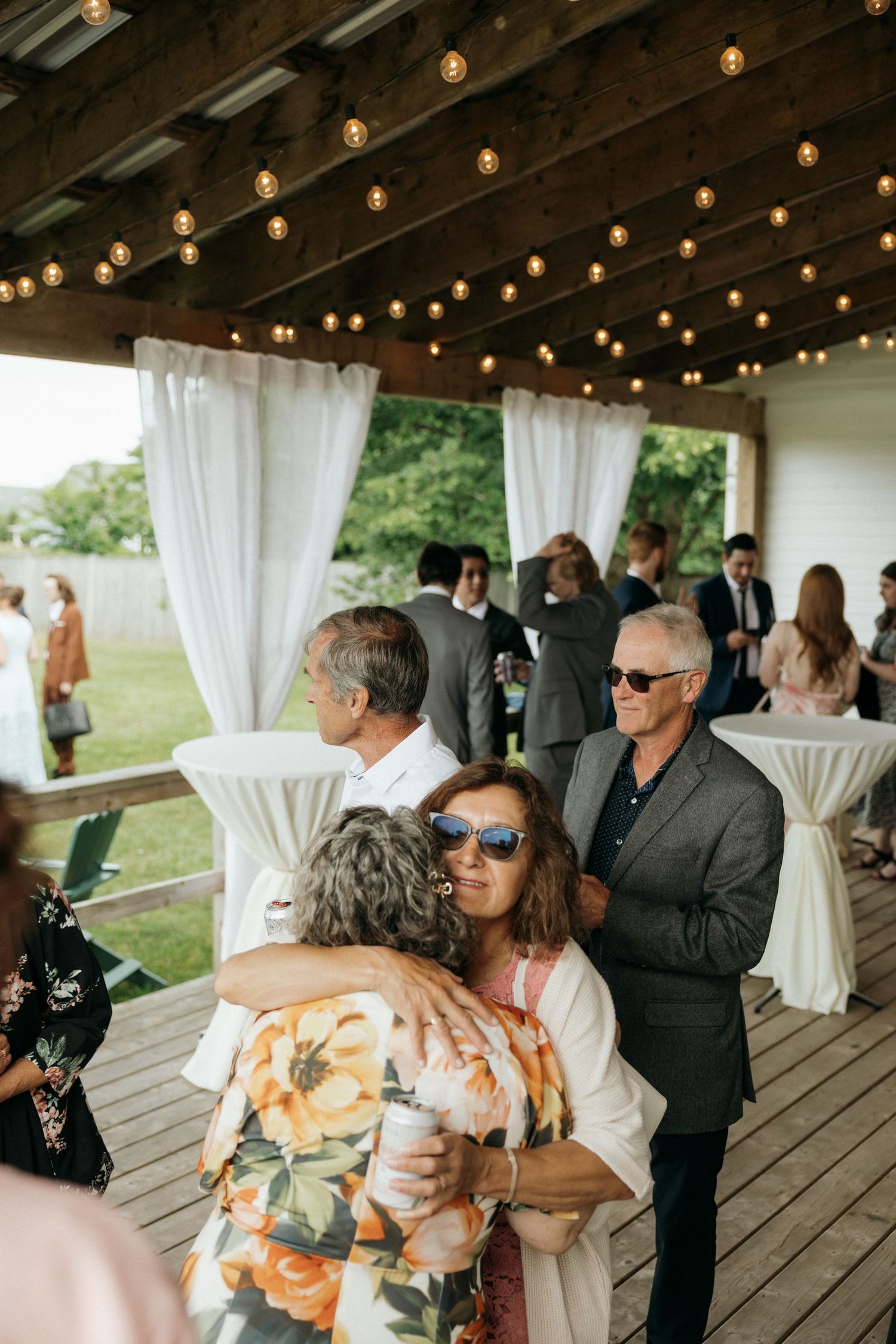 Wedding guests mingle under string lights on a covered deck during the reception.
