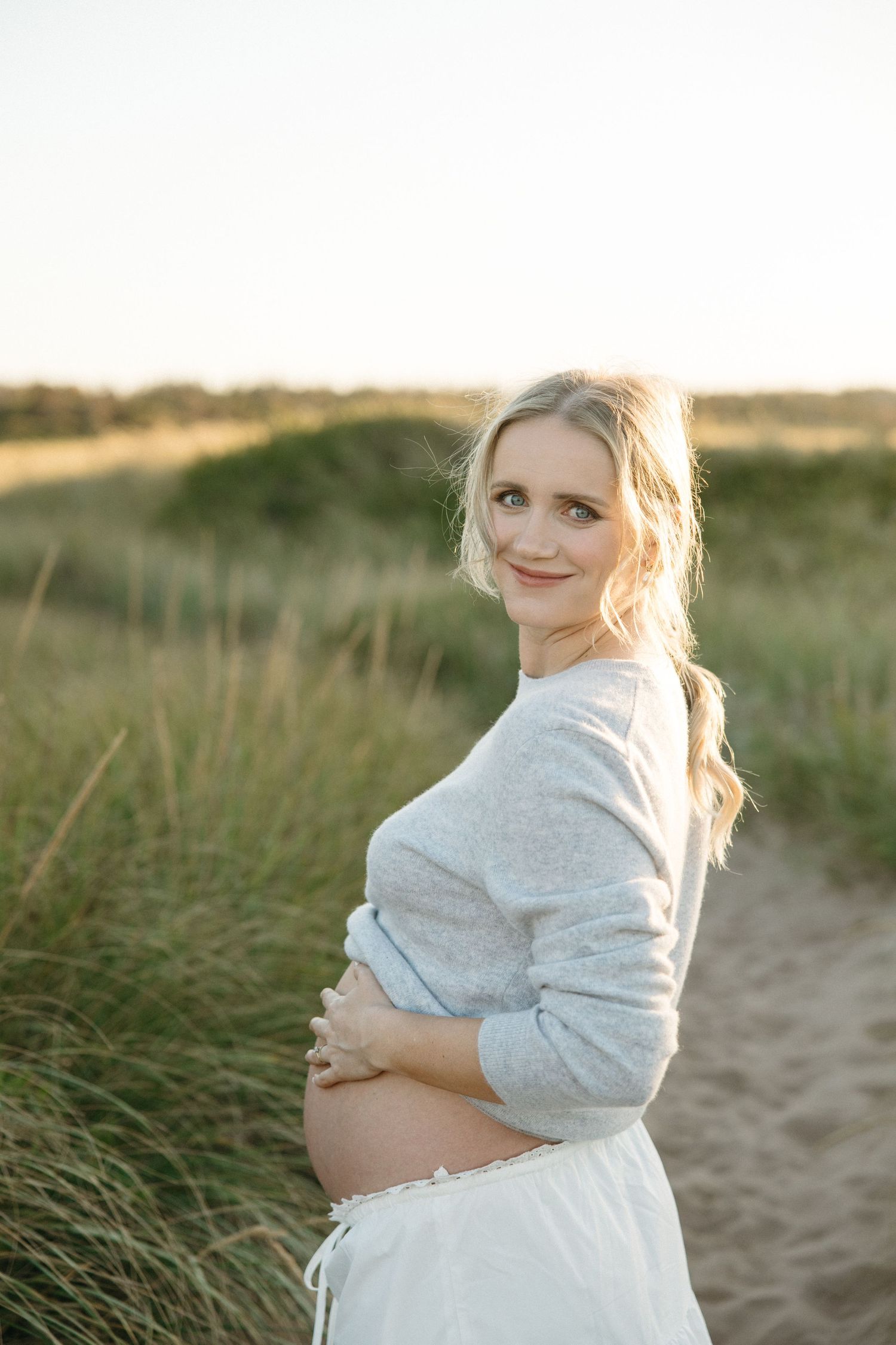 Maternity portrait in natural lighting with woman in gray sweater on sandy path.