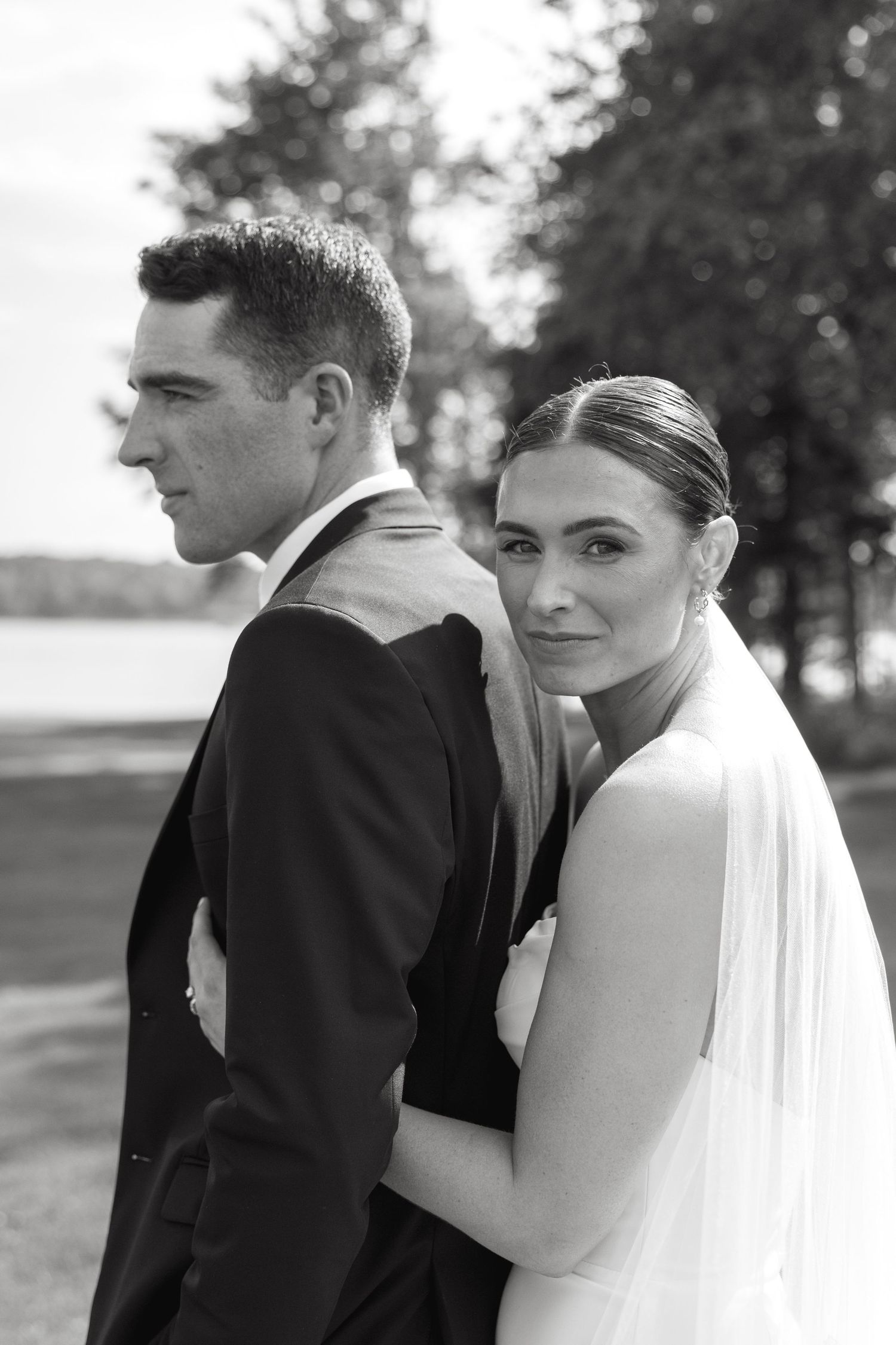 Black and white wedding portrait of bride and groom outdoors near a lake, bride smiling over shoulder.