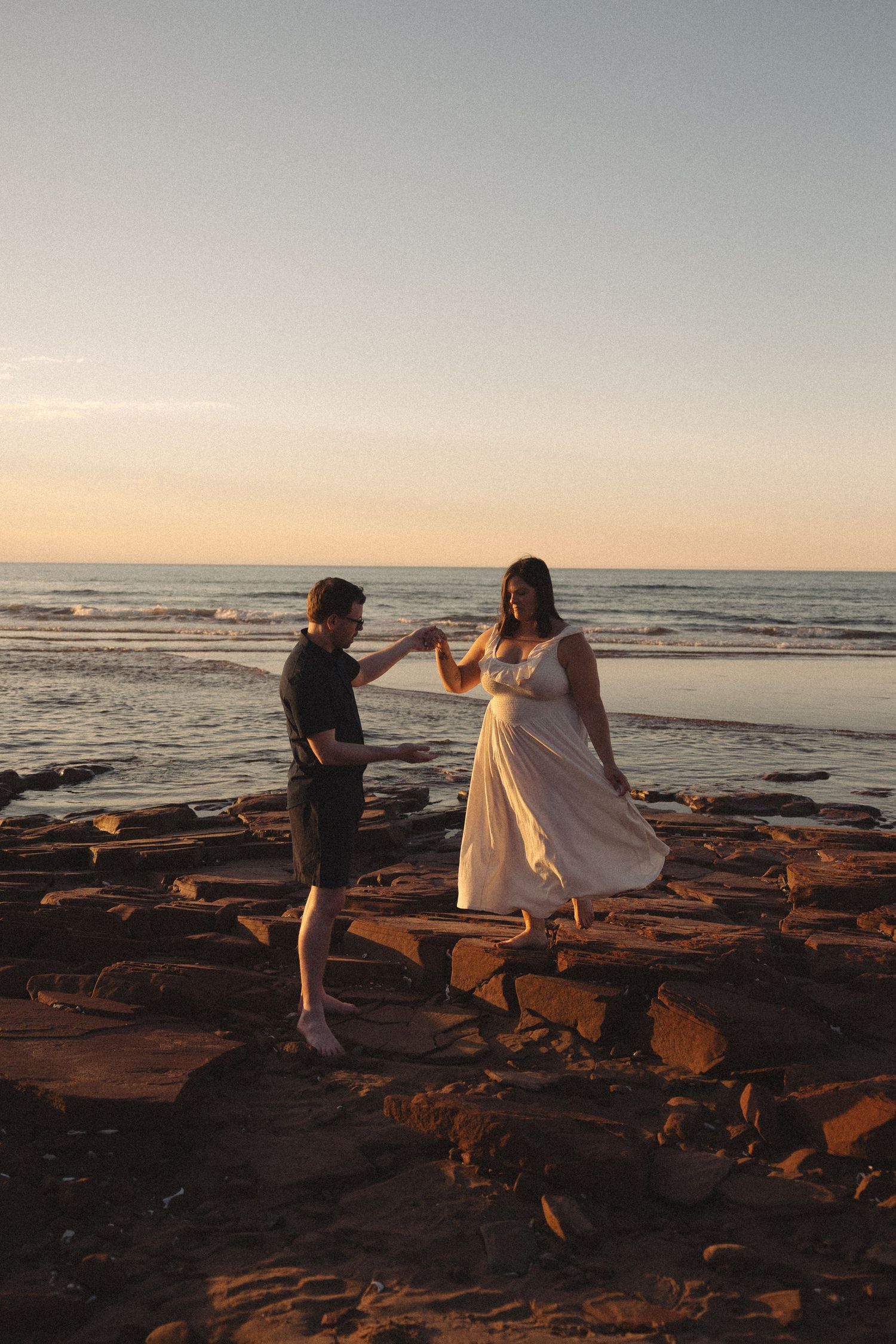 Couple dancing on rocky beach at sunset, woman in flowing white dress.