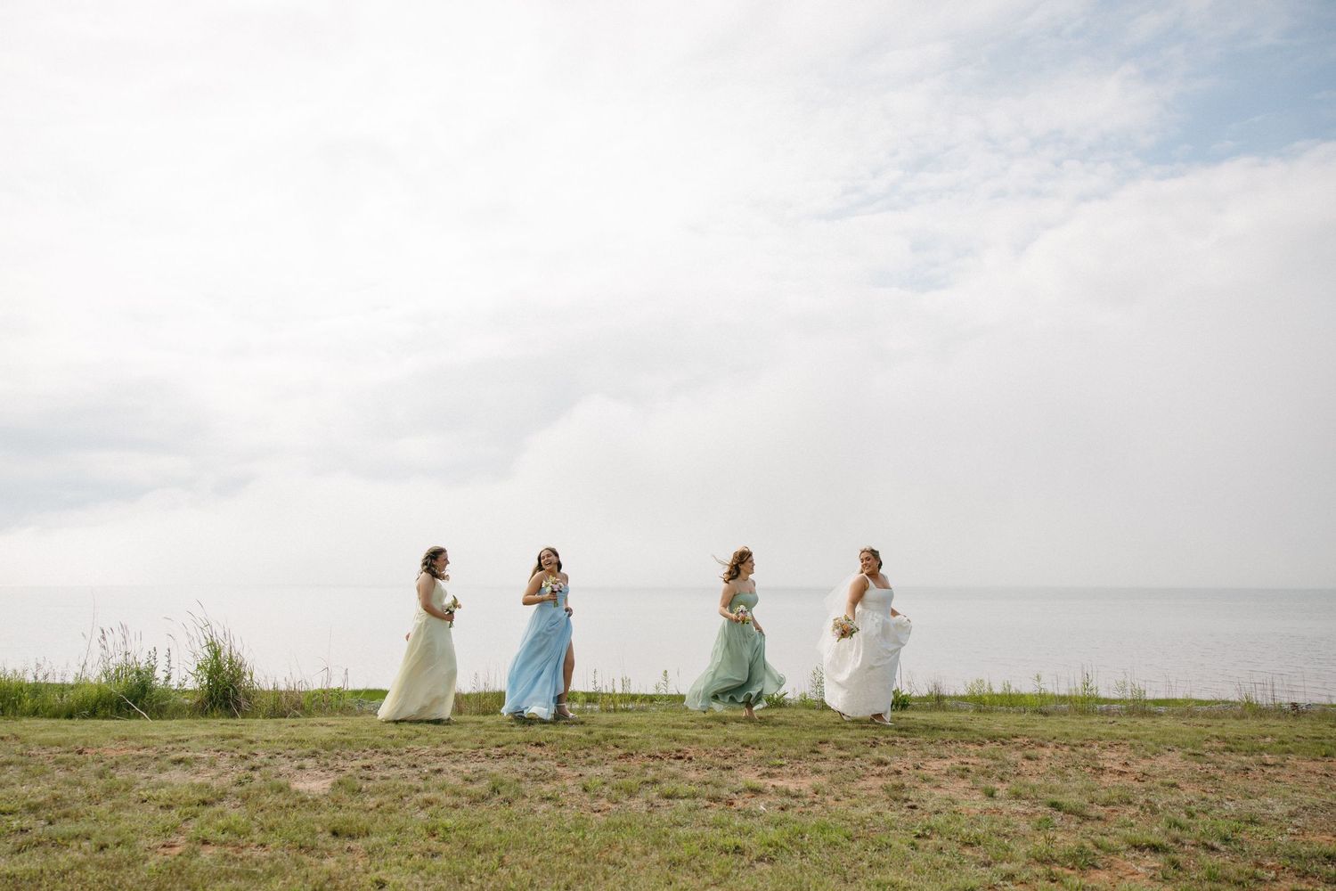 Four bridesmaids in pastel gowns walk along a grassy waterfront under a cloudy sky.