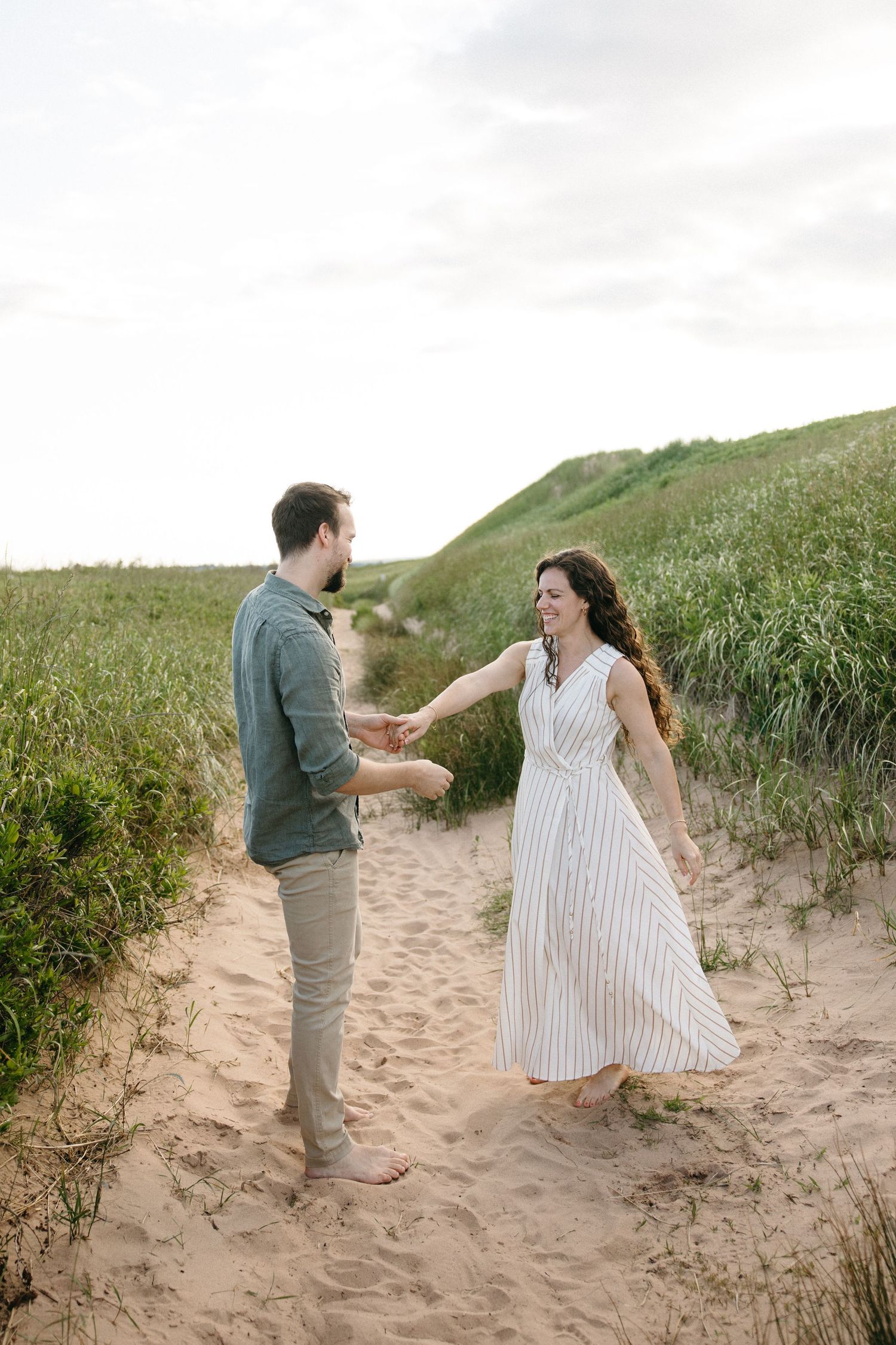 Happy couple holding hands and dancing on sandy beach path surrounded by green dune grass.
