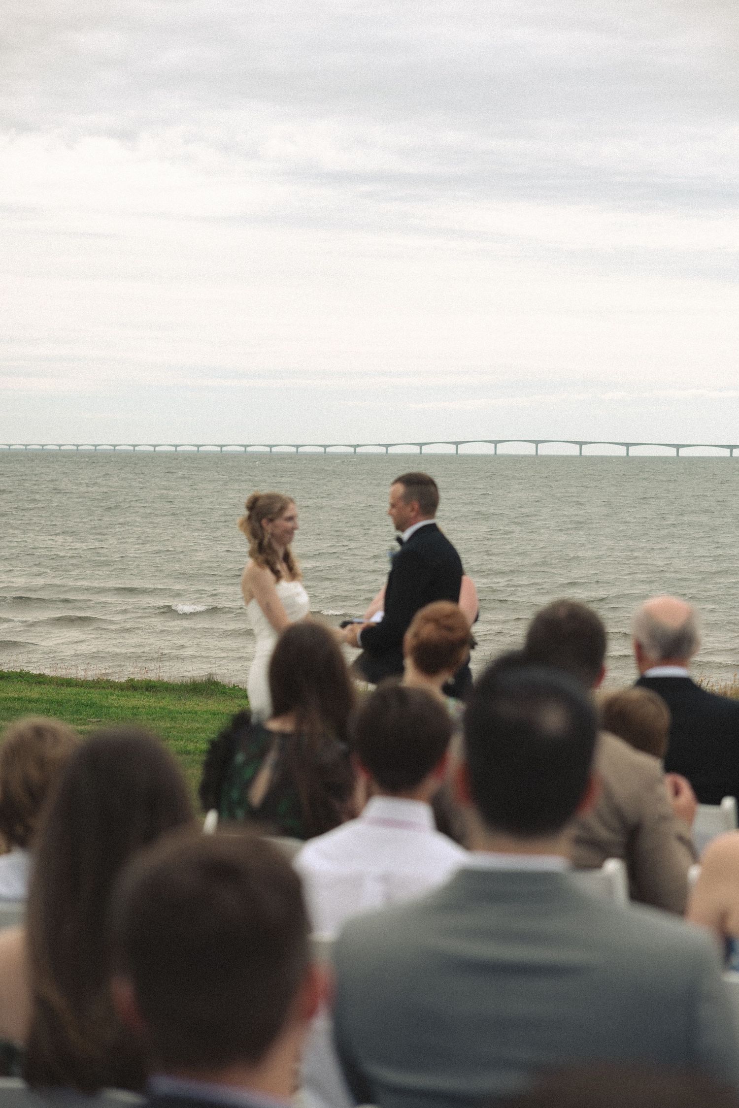 Bride and groom exchanging vows at outdoor waterfront ceremony with bridge visible over calm ocean waters.