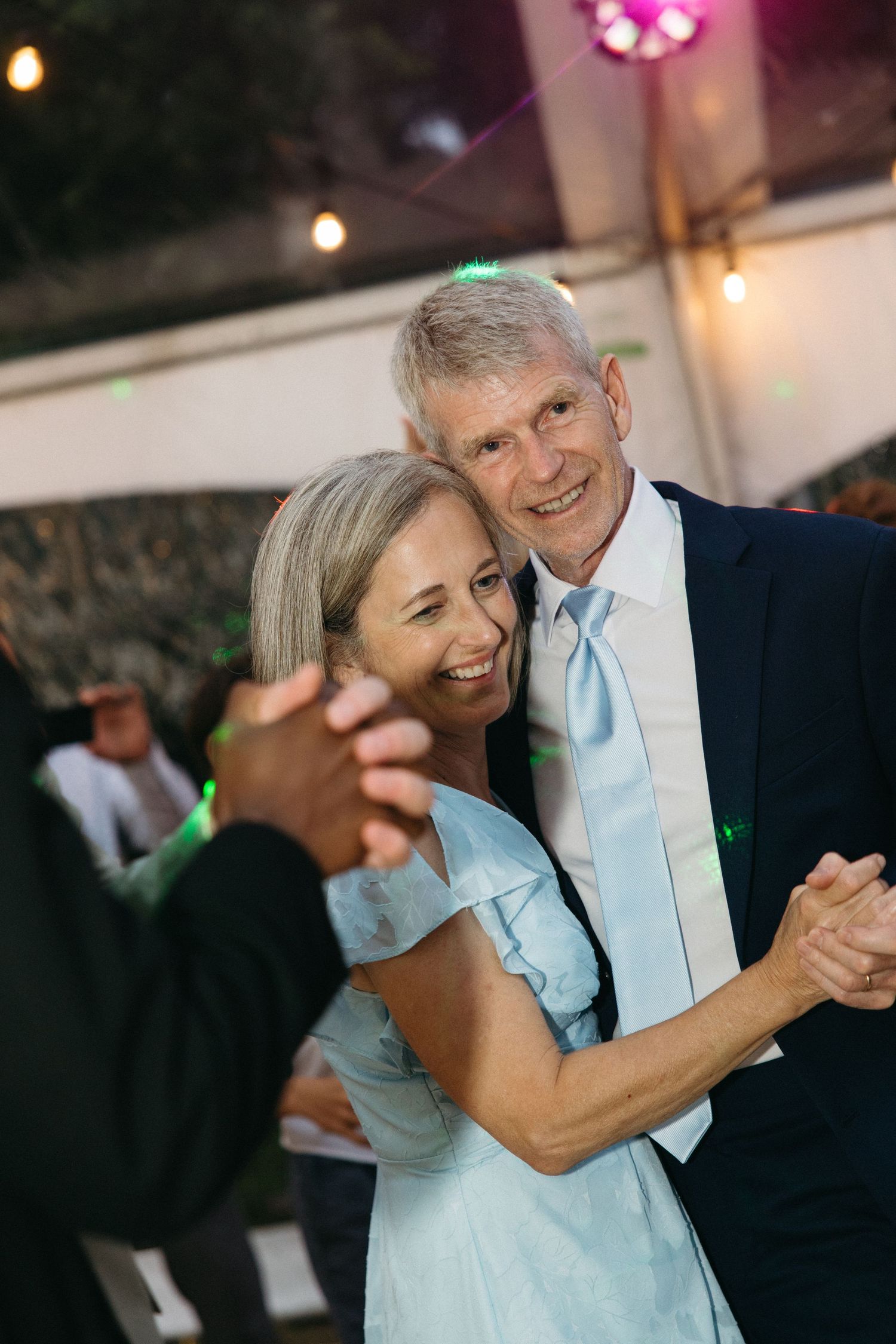 Smiling older couple dancing together at wedding reception under string lights.