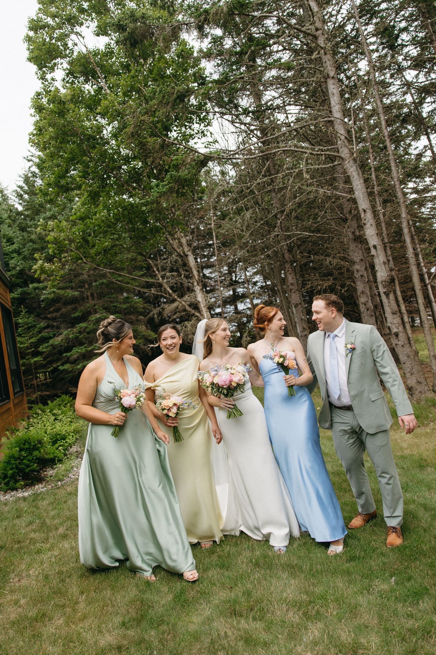 Wedding party walking outdoors with bridesmaids in sage green, gold, and blue satin gowns holding bouquets.