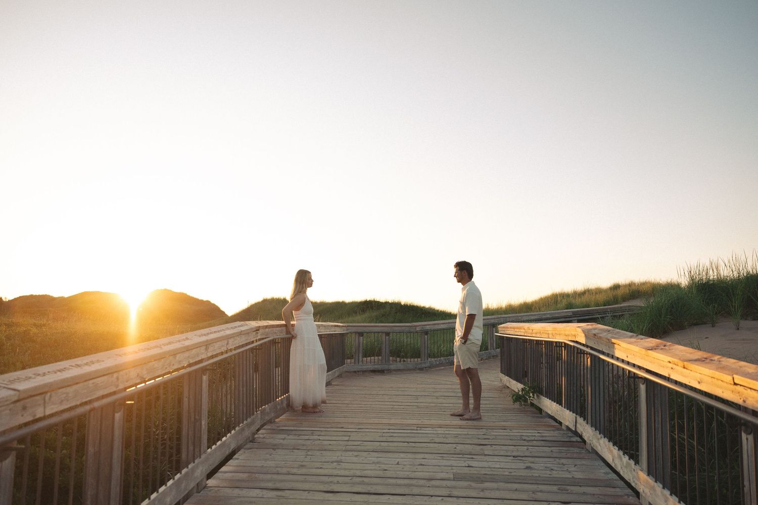 Couple facing each other on a wooden boardwalk at golden sunset near sand dunes and beach grass.