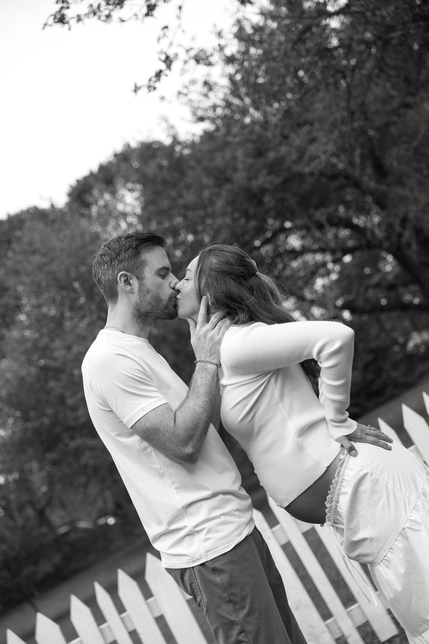 Black and white photo of a couple sharing a passionate kiss beside a white picket fence outdoors.