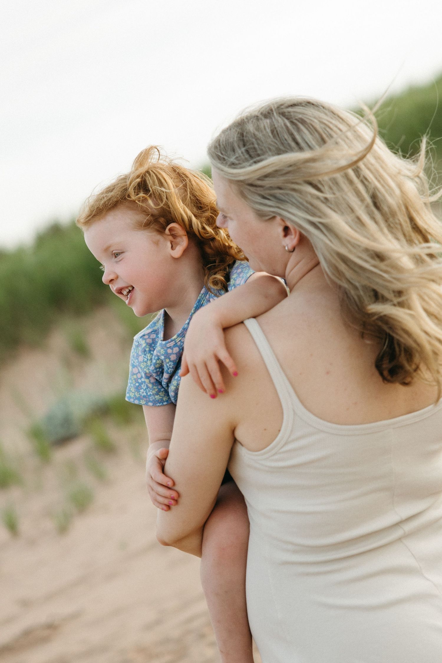 Blonde mother carrying laughing red-haired daughter on her back at the beach.