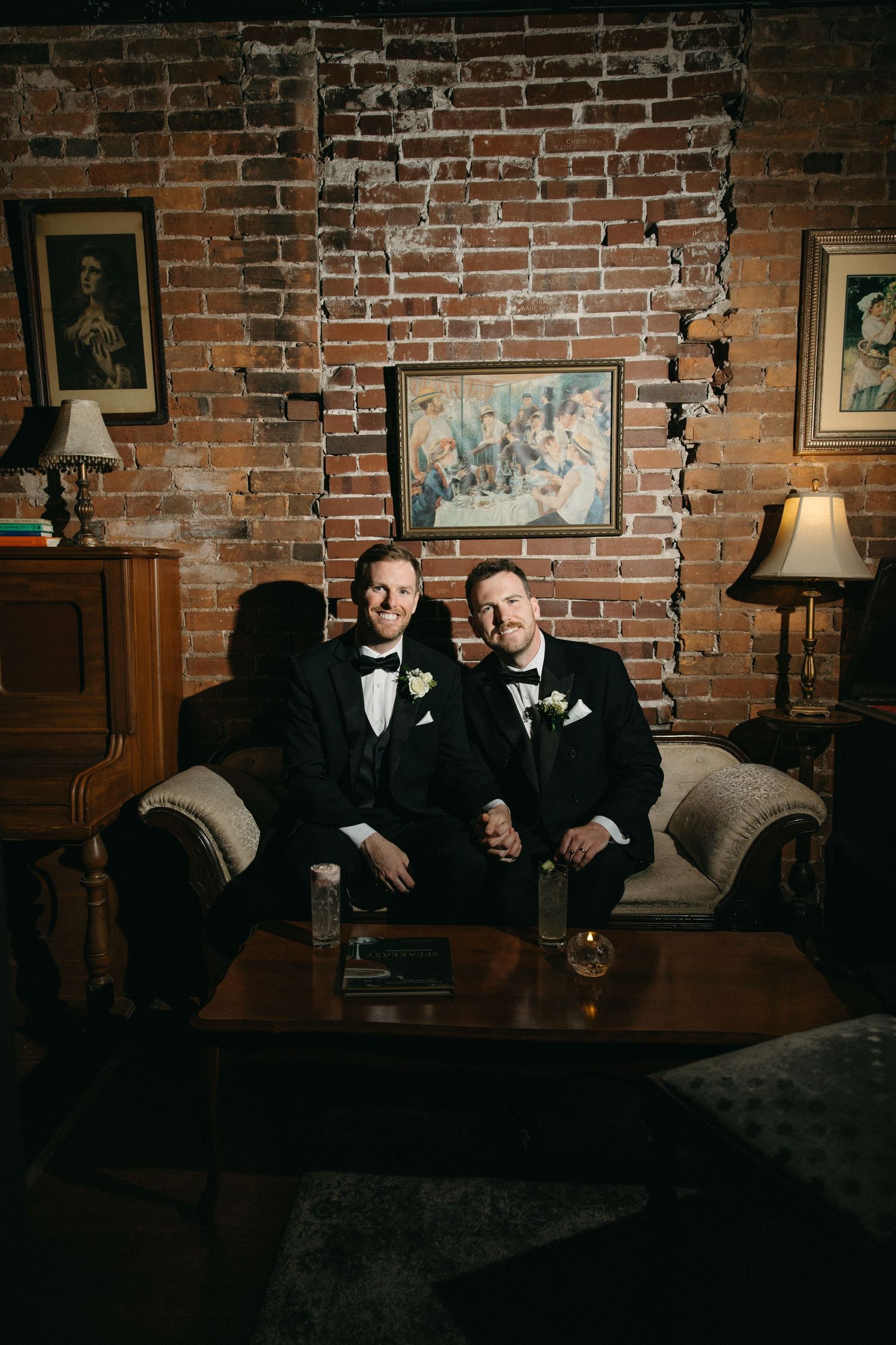 Two people in formal black suits relax on a vintage couch against exposed brick wall with framed artwork.