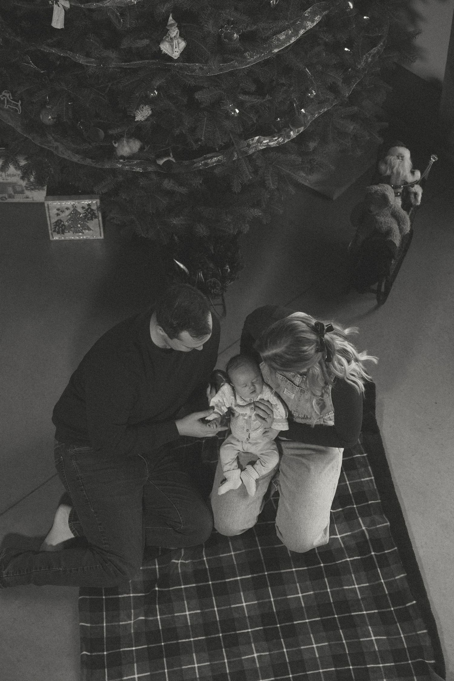 A family gathers on plaid blanket beneath decorated Christmas tree in dark room.