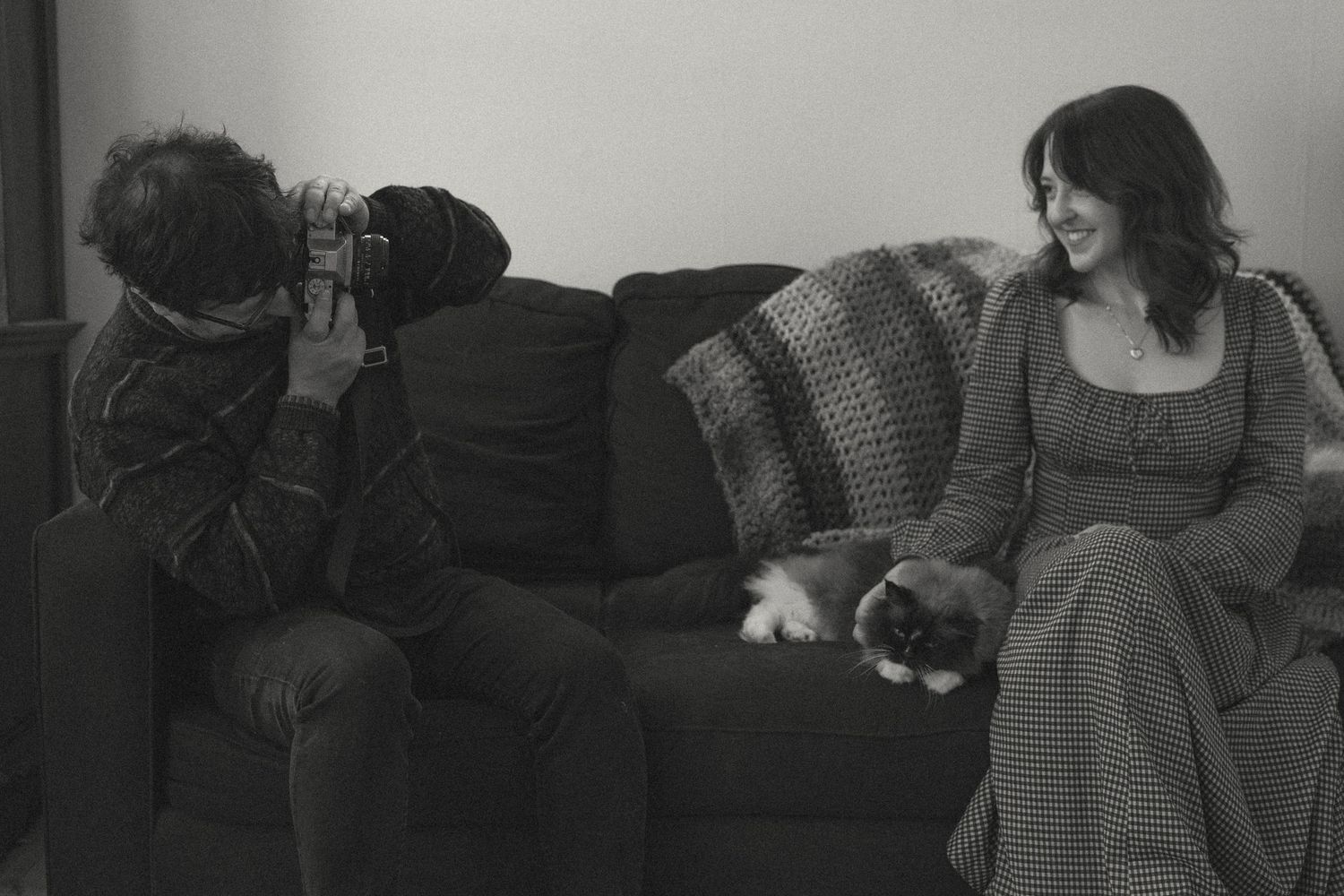 Black and white photo of a photographer taking pictures of a smiling woman holding a cat on a couch.