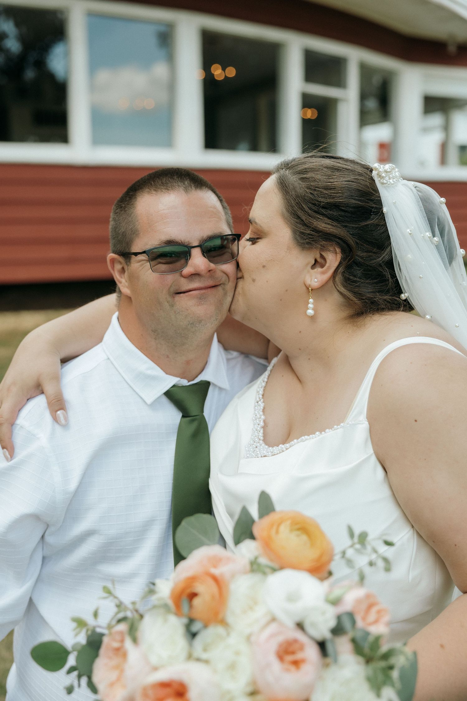 Bride in white gown kissing groom on cheek while holding a peach and white bouquet near a red barn.
