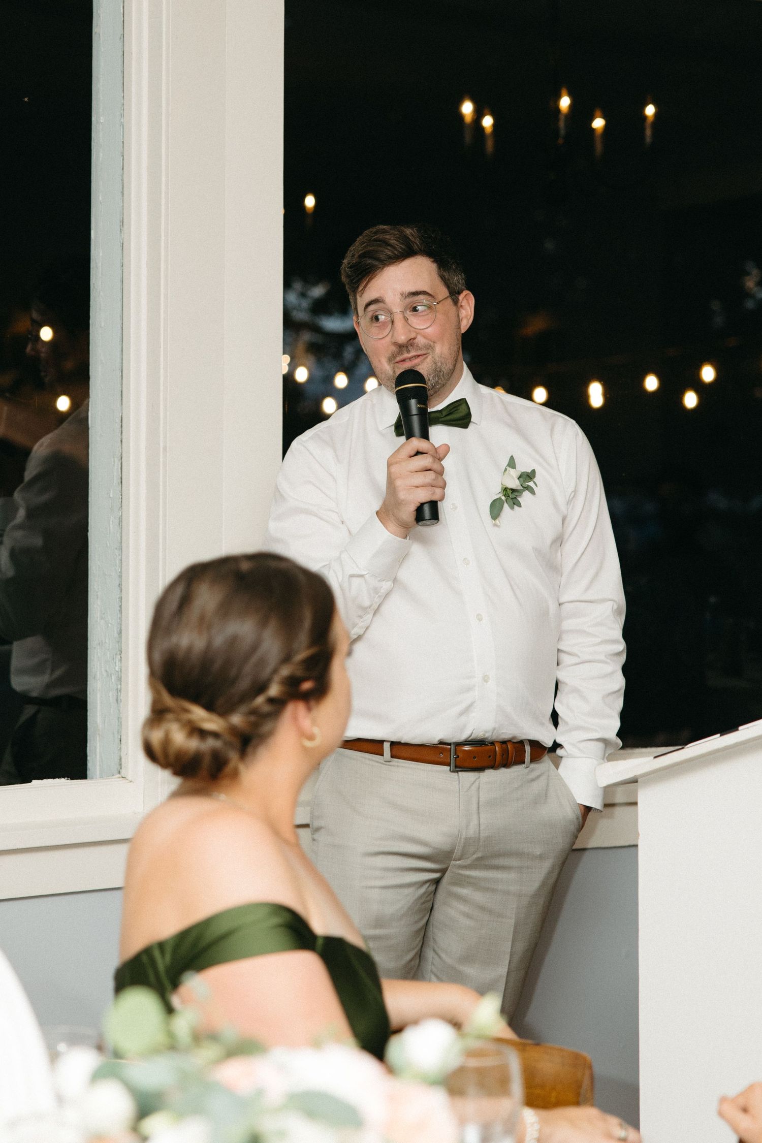 Best man in white shirt and green bow tie gives a speech at indoor wedding reception.