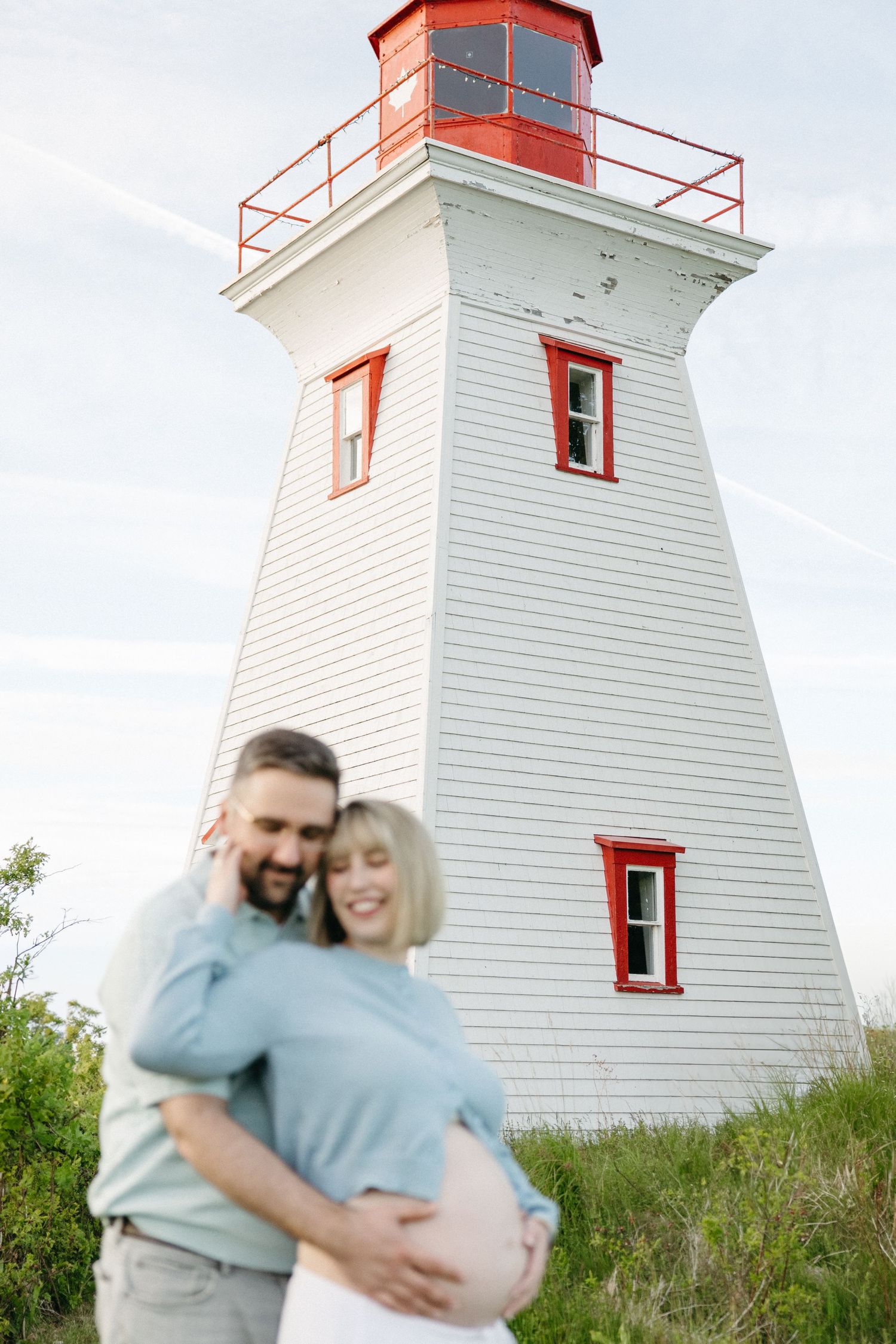 Happy couple embracing pregnant belly in front of white and red lighthouse on sunny day.