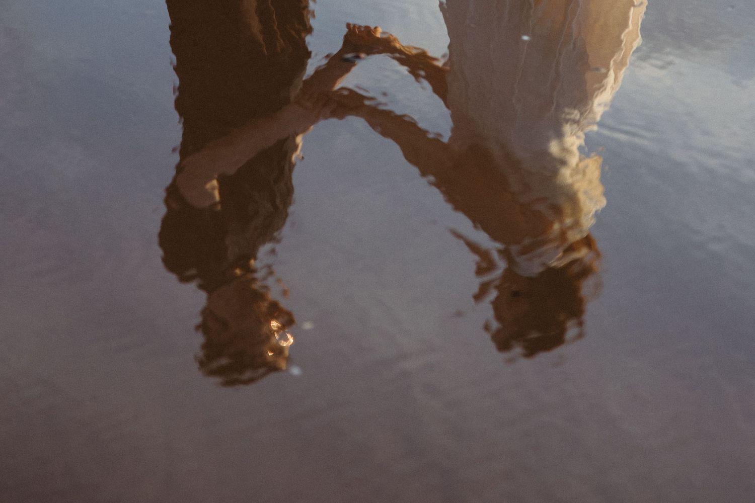 Reflection of two people holding hands in shallow water on a beach at golden hour.