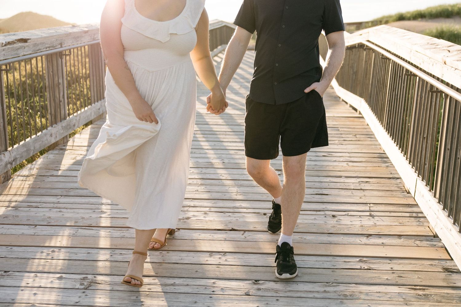 Couple holding hands strolling on sun-drenched wooden boardwalk near beach at golden hour.