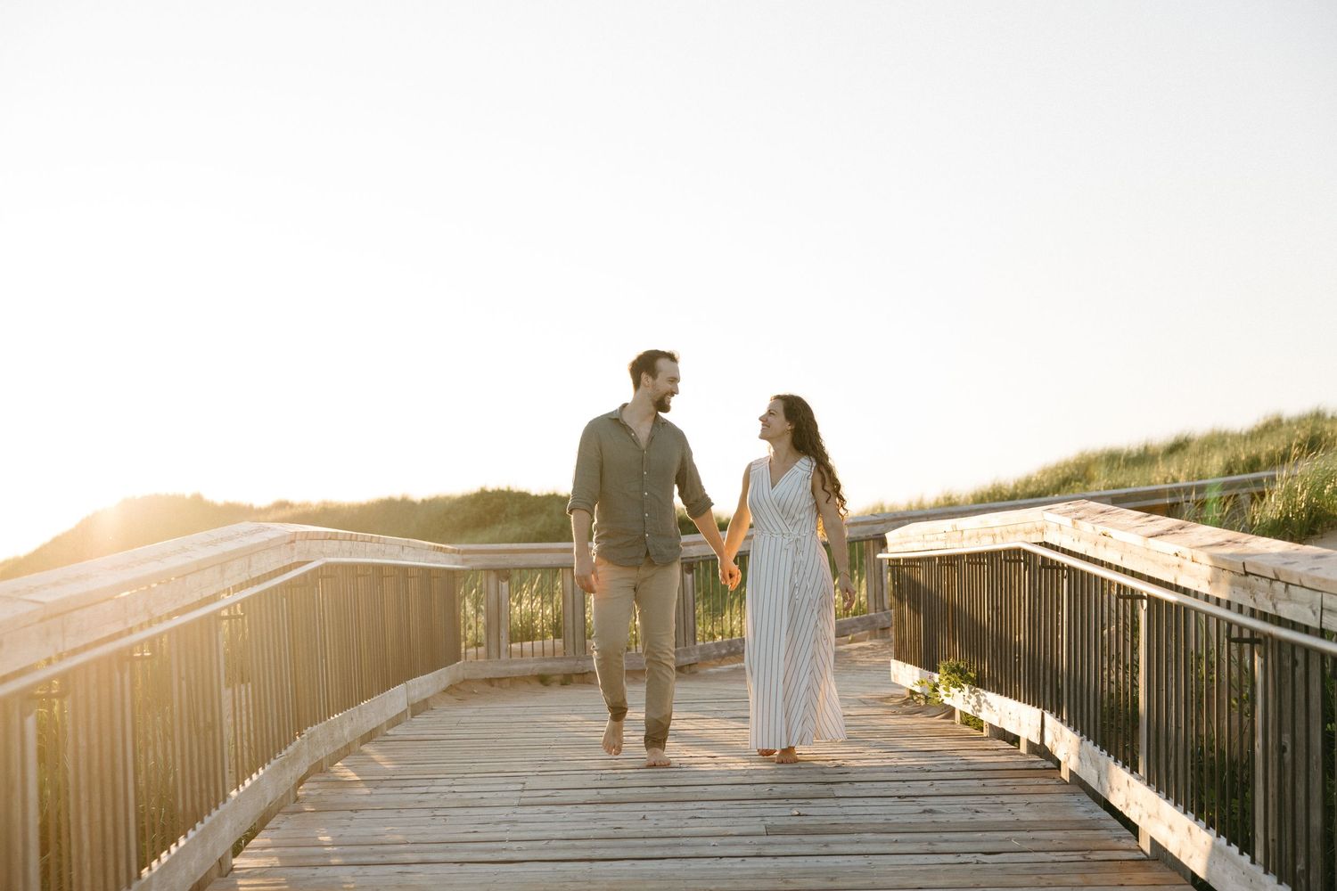Couple holding hands walking on a sunny wooden boardwalk surrounded by dunes and golden light.