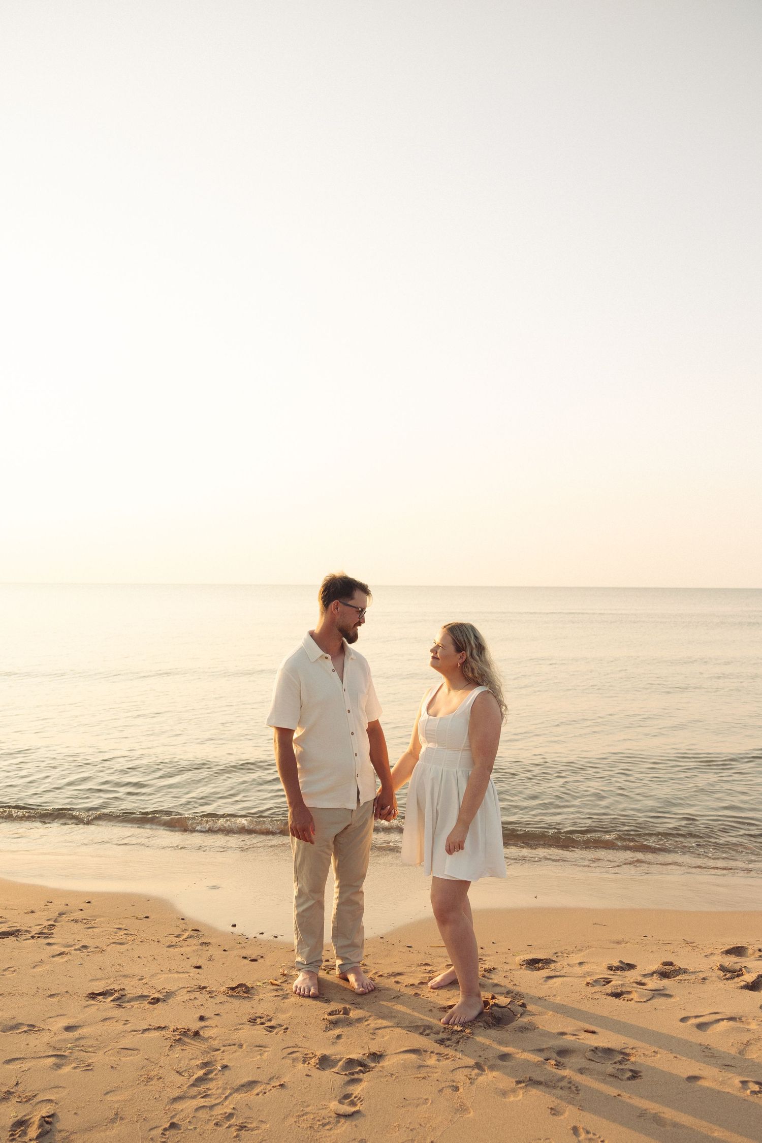 Couple holding hands on a sandy beach at sunset, wearing white outfits, with calm ocean waves in the background.