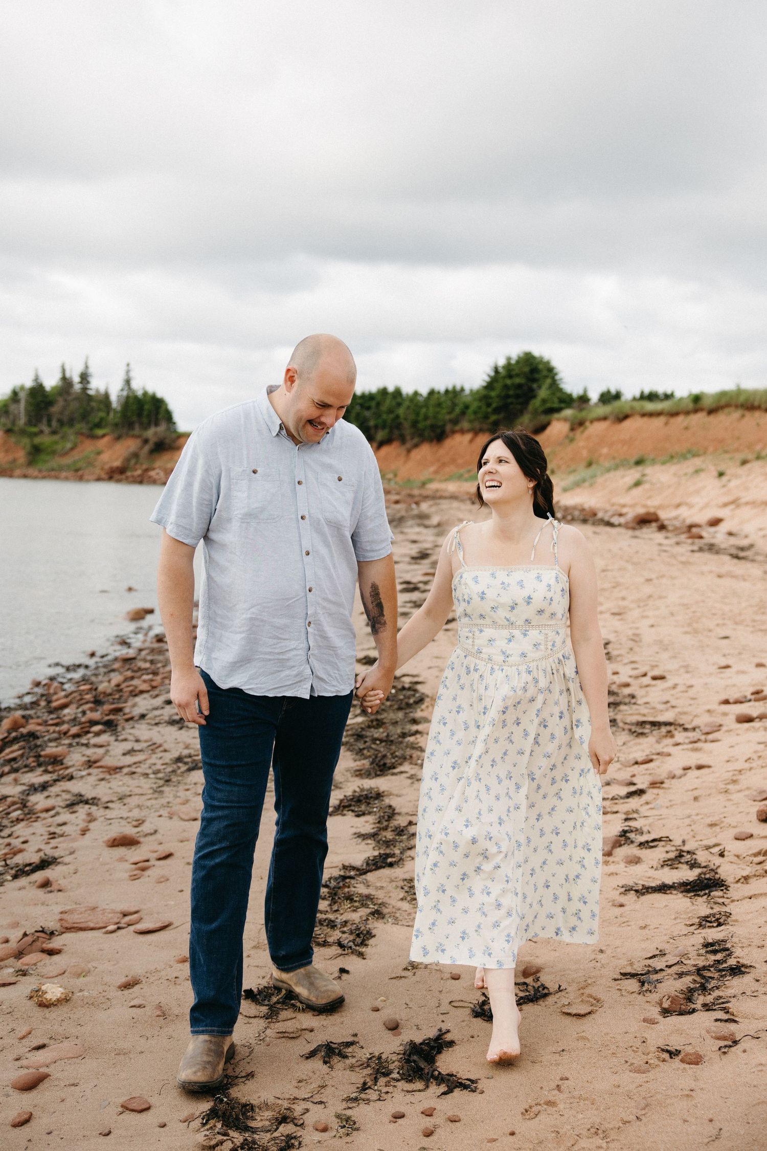 Happy couple holding hands and laughing while walking along a rocky red sand beach shoreline.