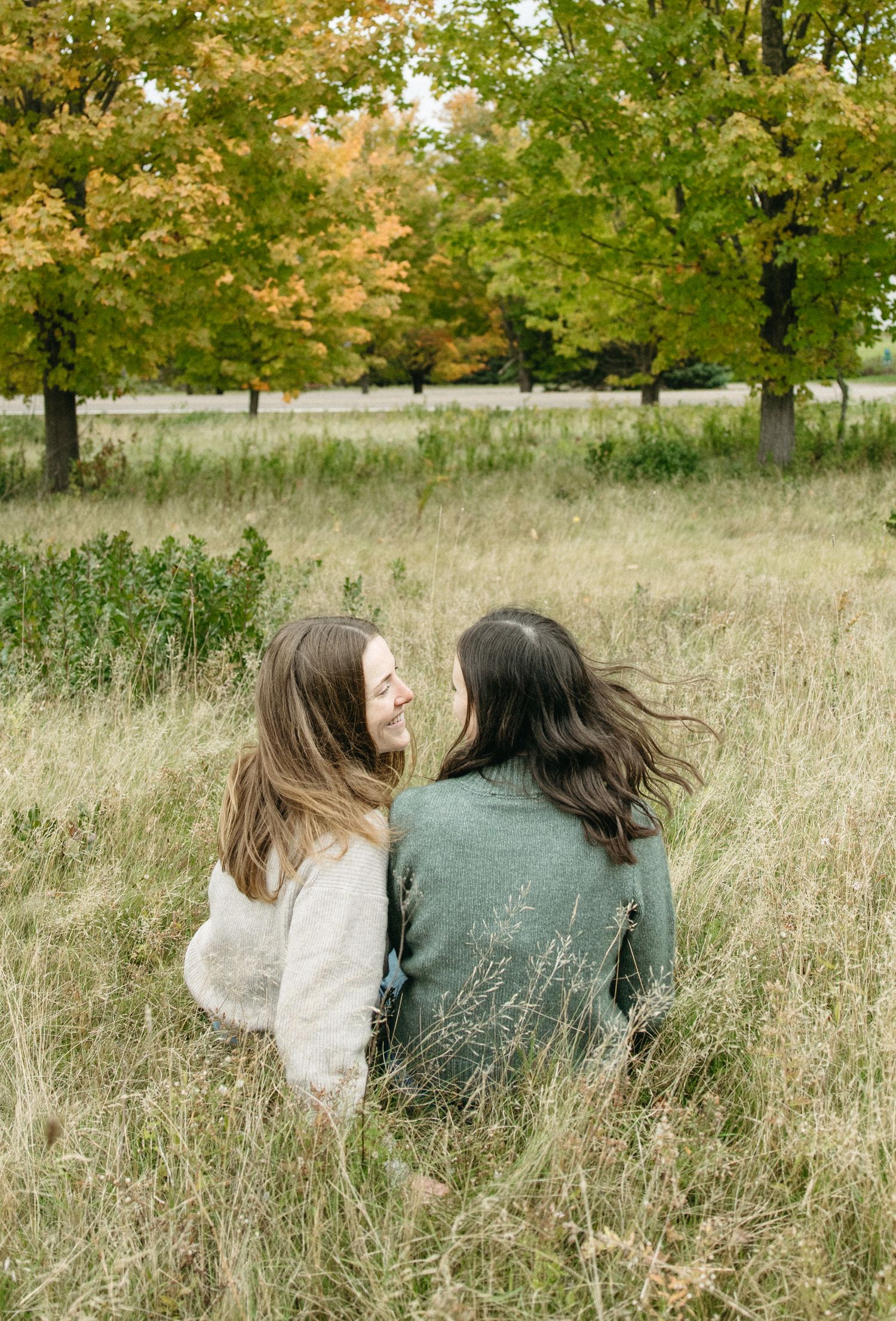 Two women in cozy sweaters sit side by side in tall golden grass, facing each other near autumn trees.