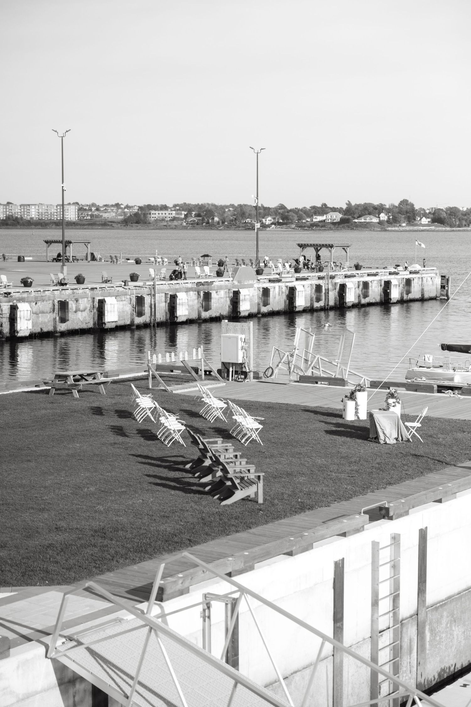 Black and white view of a waterfront lawn with folding chairs and a table beside a harbor dock, with lamp posts and a distant shoreline.