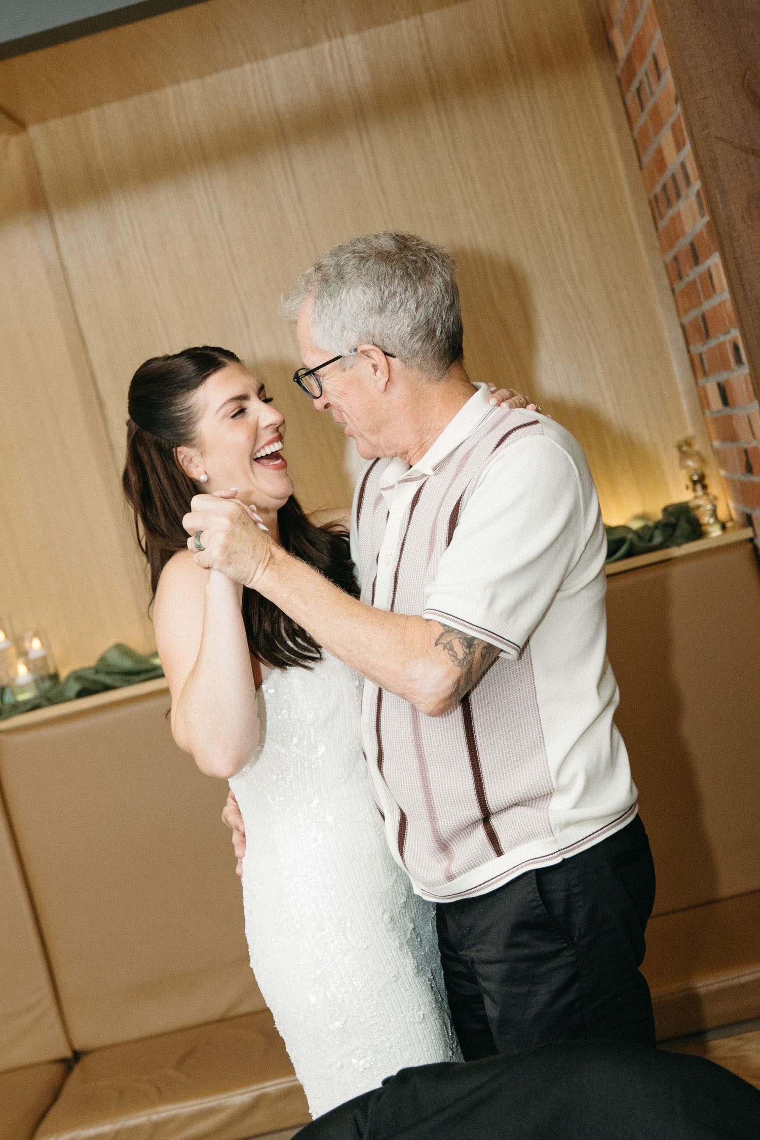 A smiling bride in a white lace gown laughs while dancing with an older gray-haired man in glasses at a wedding reception.