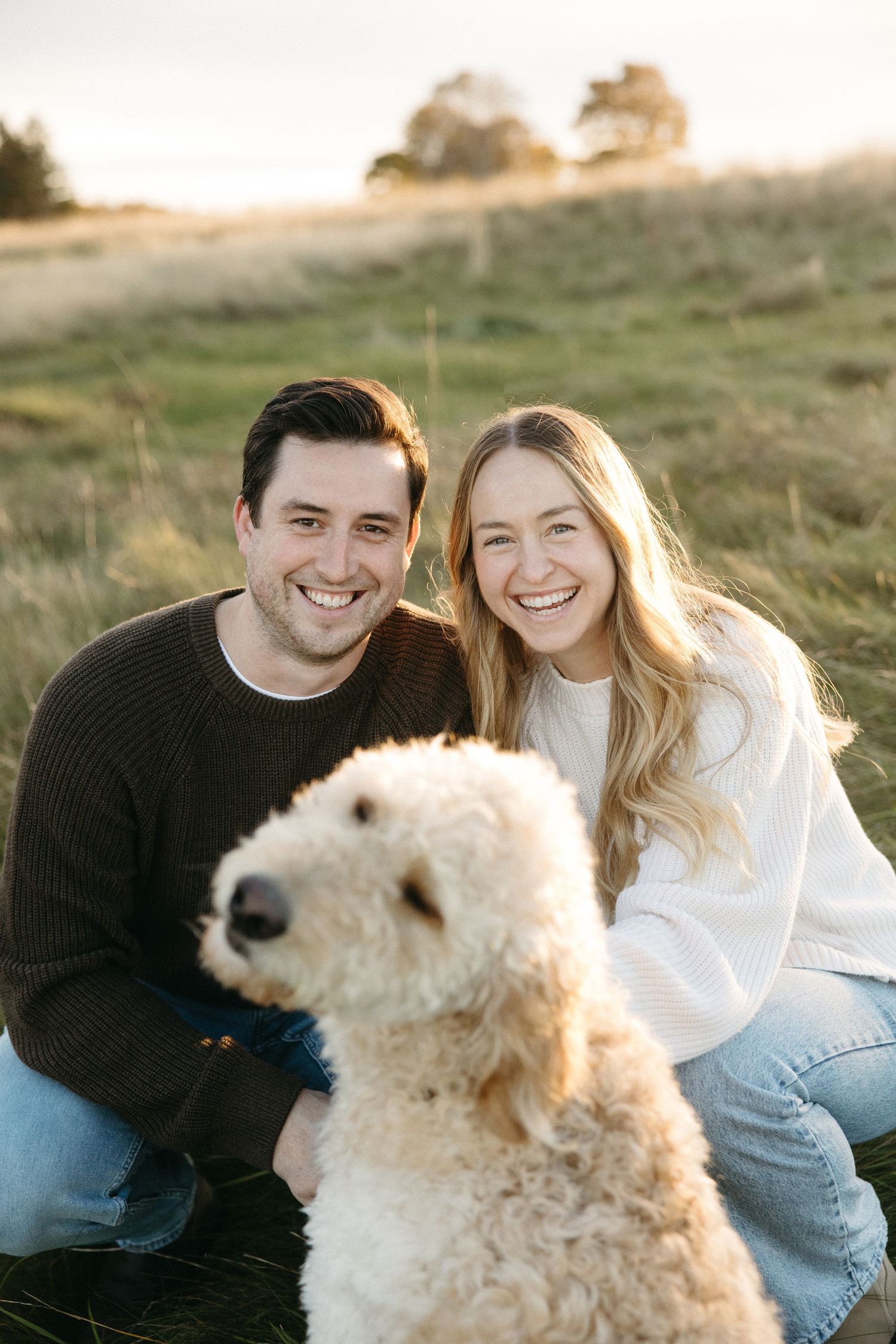 A smiling couple kneels in a grassy field at sunset with their fluffy cream goldendoodle in the foreground.
