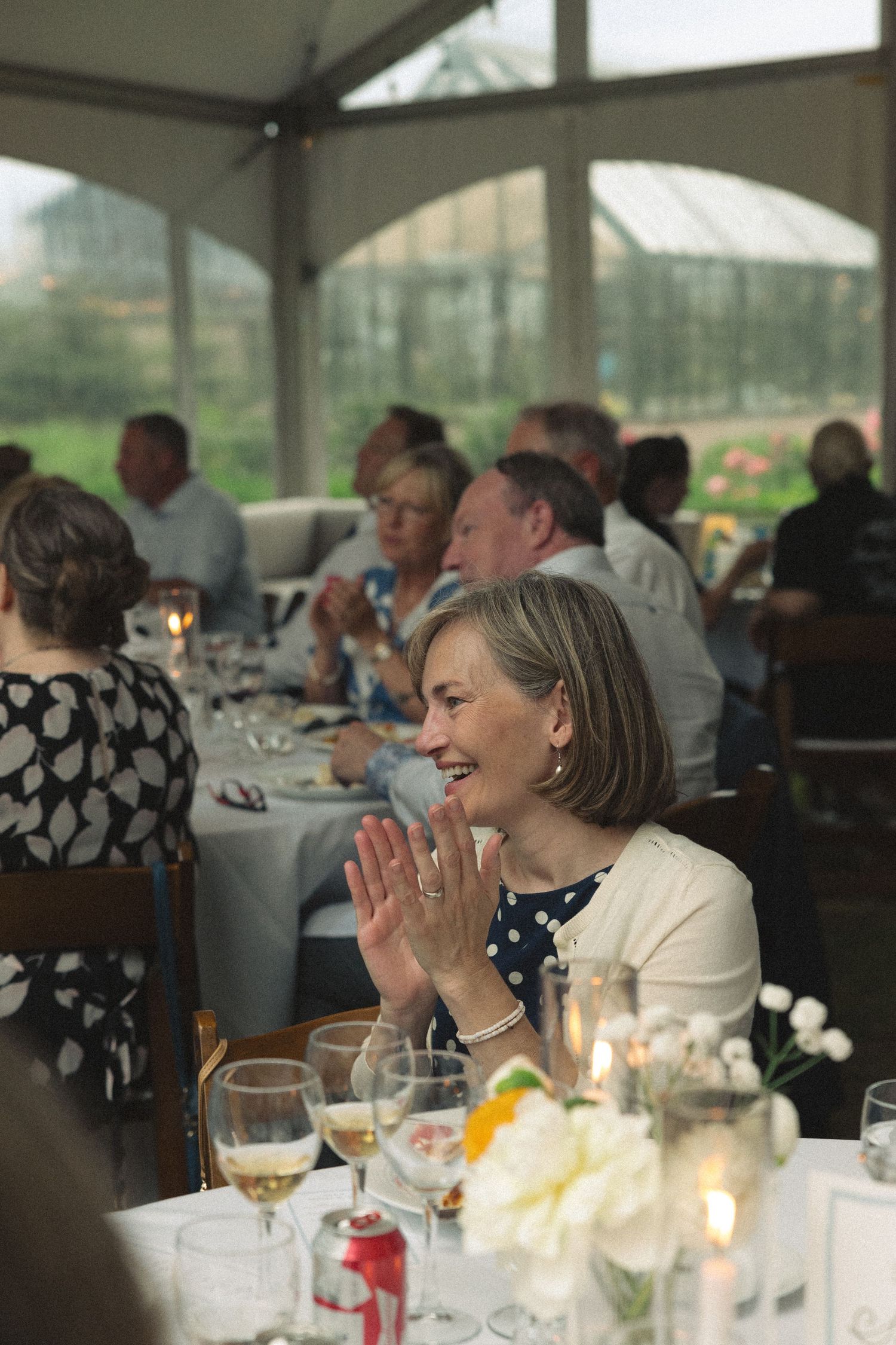 Smiling woman in white blazer clapping at elegant wedding reception dinner under a tent with guests.