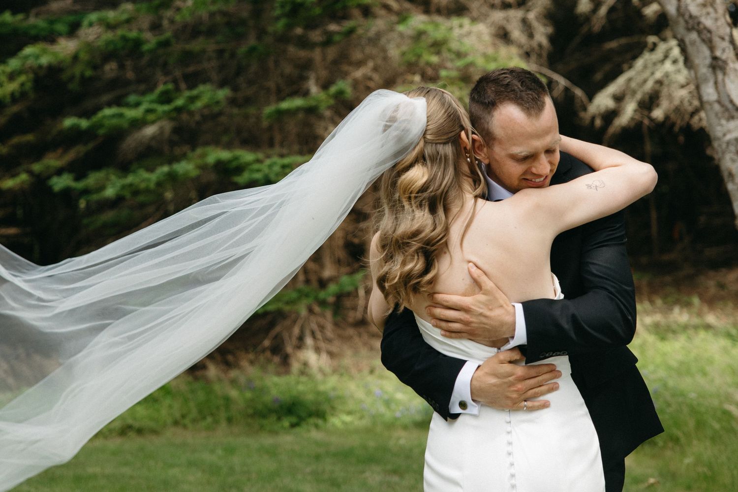 Bride and groom embracing outdoors with veil flowing in wind, surrounded by lush greenery on their wedding day.