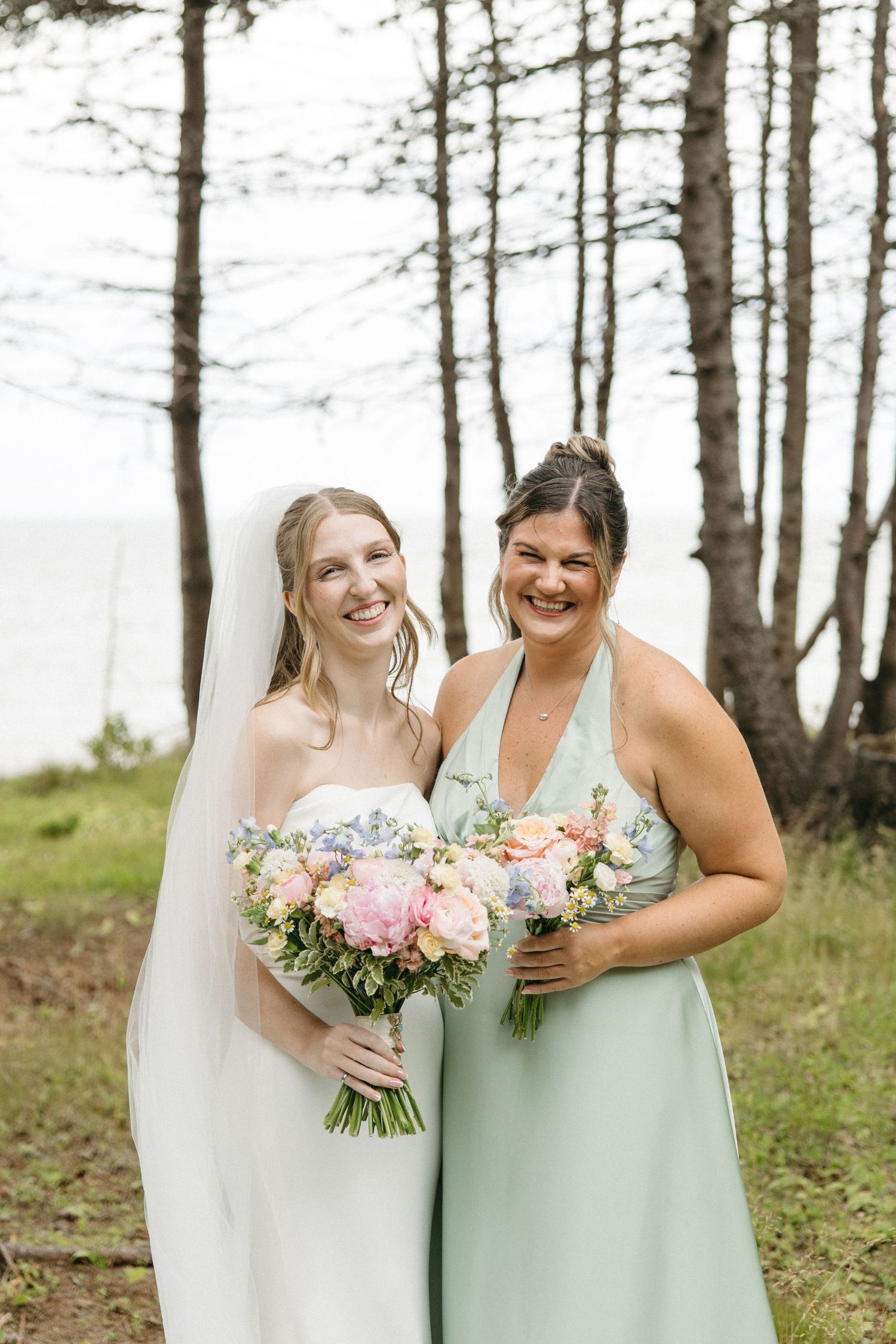 Bride in white gown and bridesmaid in sage green dress holding pastel bouquets, smiling outdoors among pine trees.