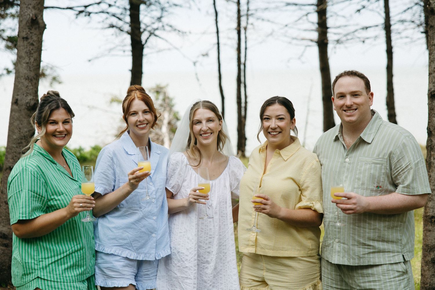 Five friends smiling outdoors among trees holding orange drinks wearing colorful striped pajamas at a casual gathering.