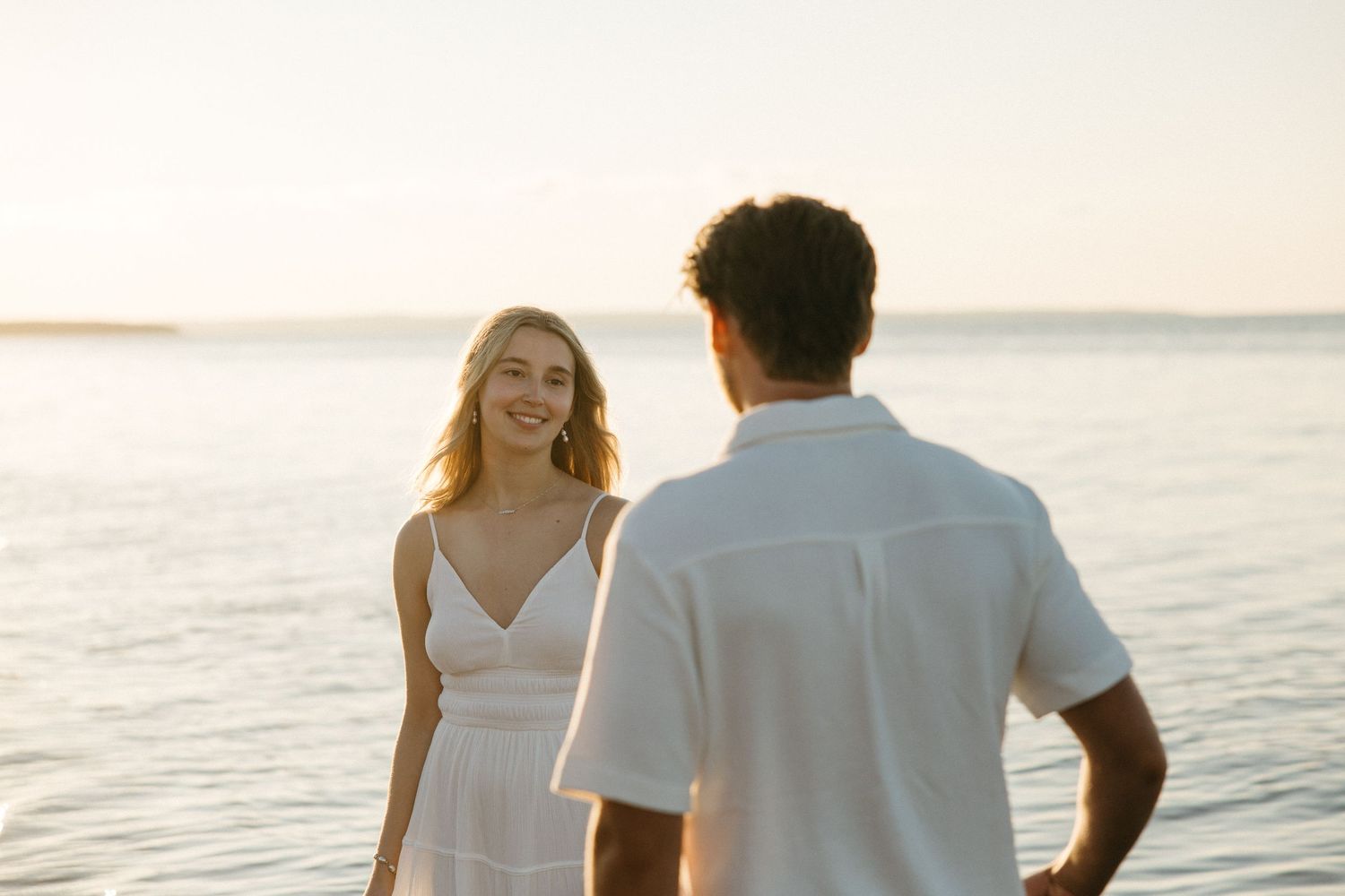Smiling blonde woman in white dress facing man near ocean at sunset.