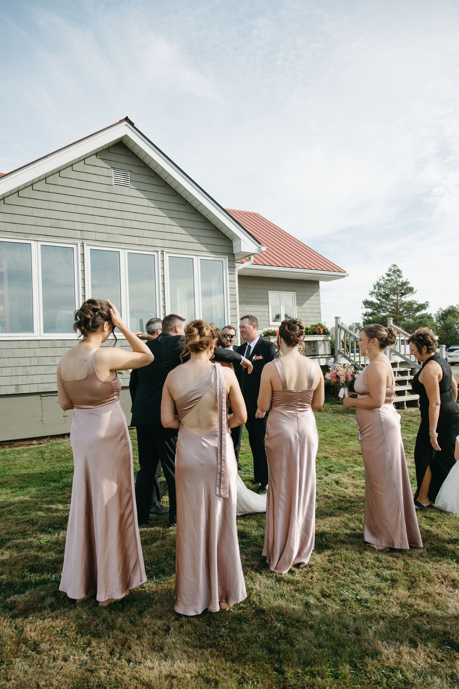 Wedding party in pink dresses stands on a grassy hill beside a white building.