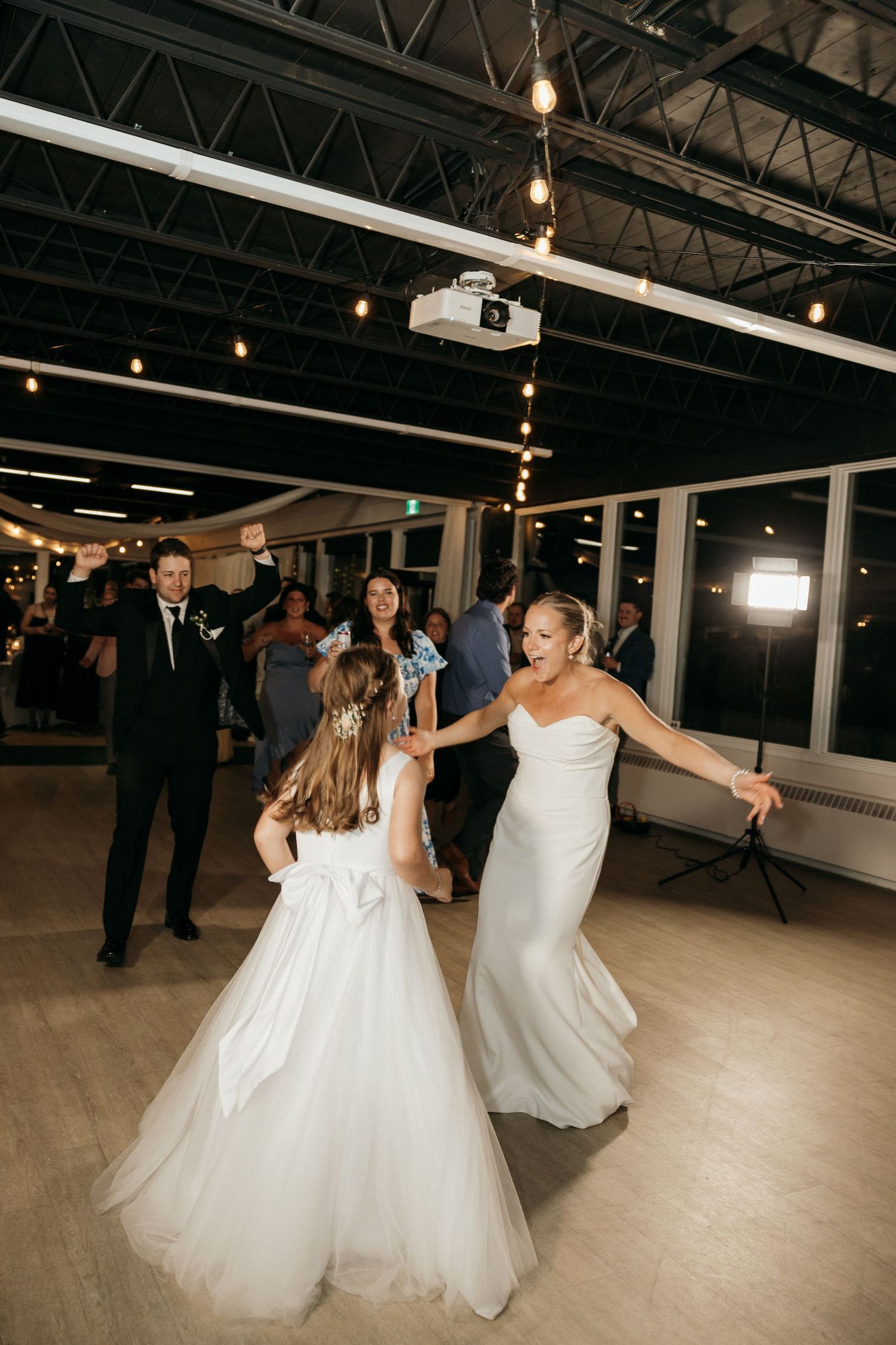Two people in white dresses twirl and dance together on a spacious dance floor with industrial ceiling.