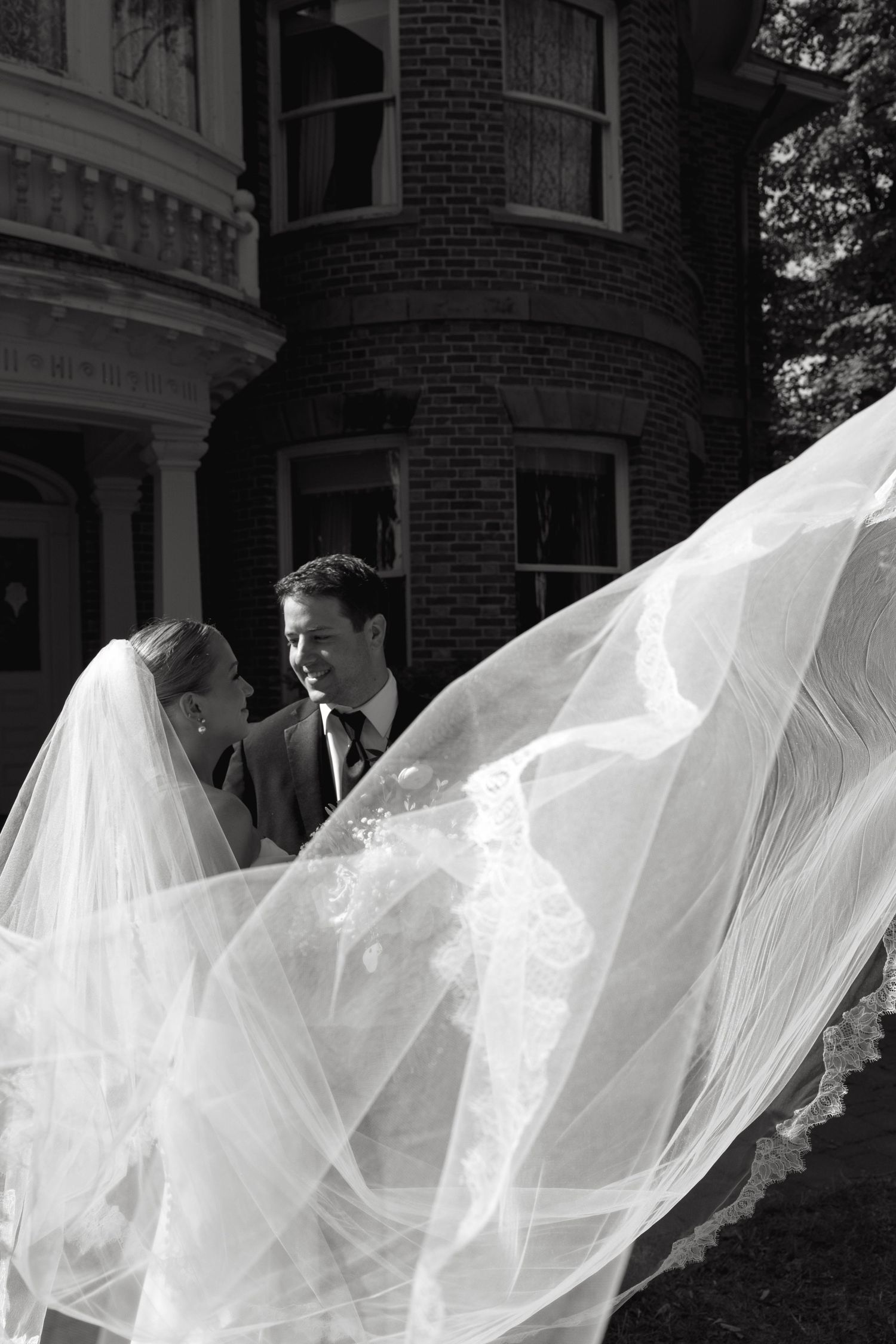 Black and white portrait with flowing veil against brick building exterior.
