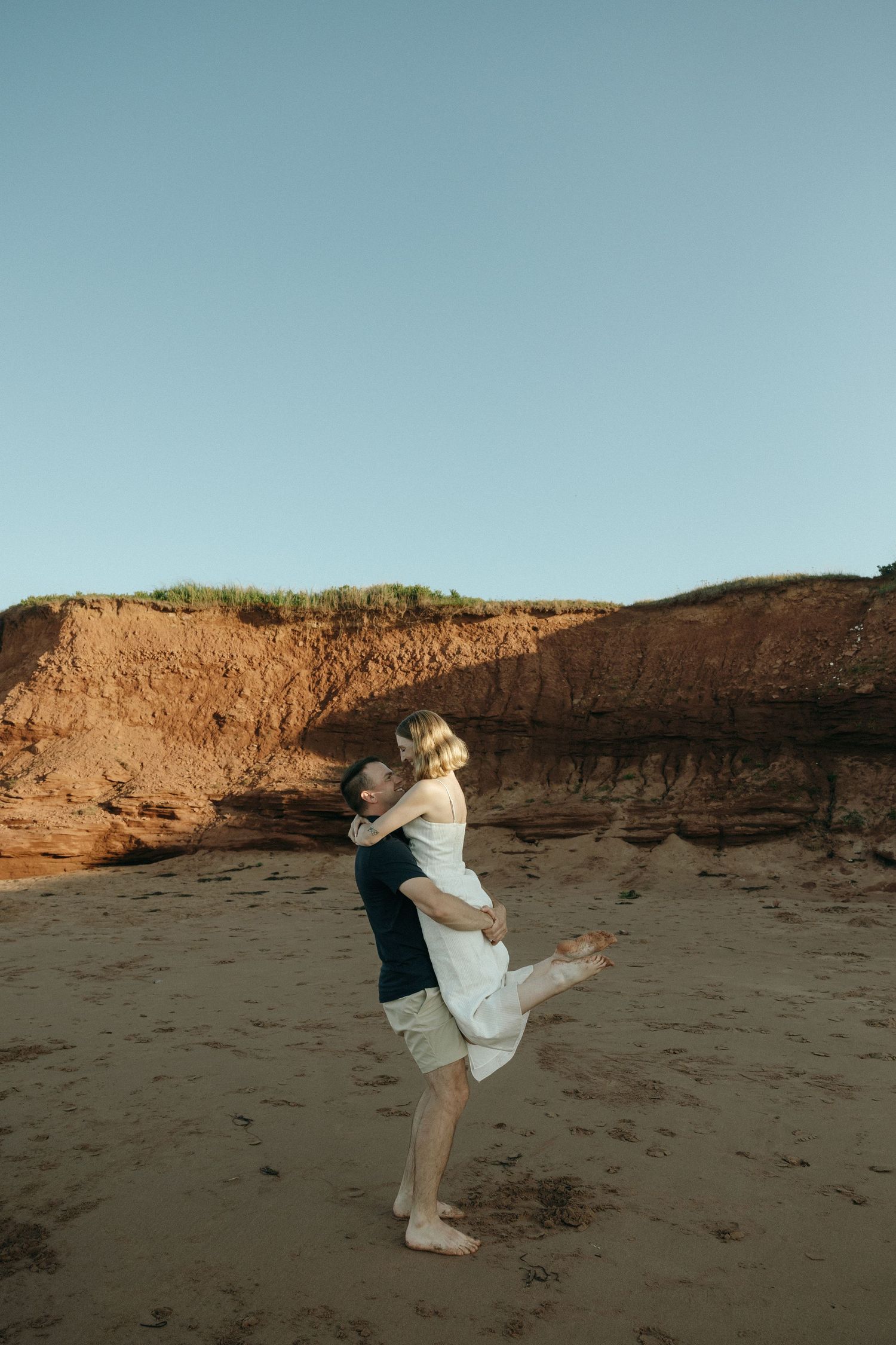 Playful couple dancing and spinning on beach with cliffs in background.