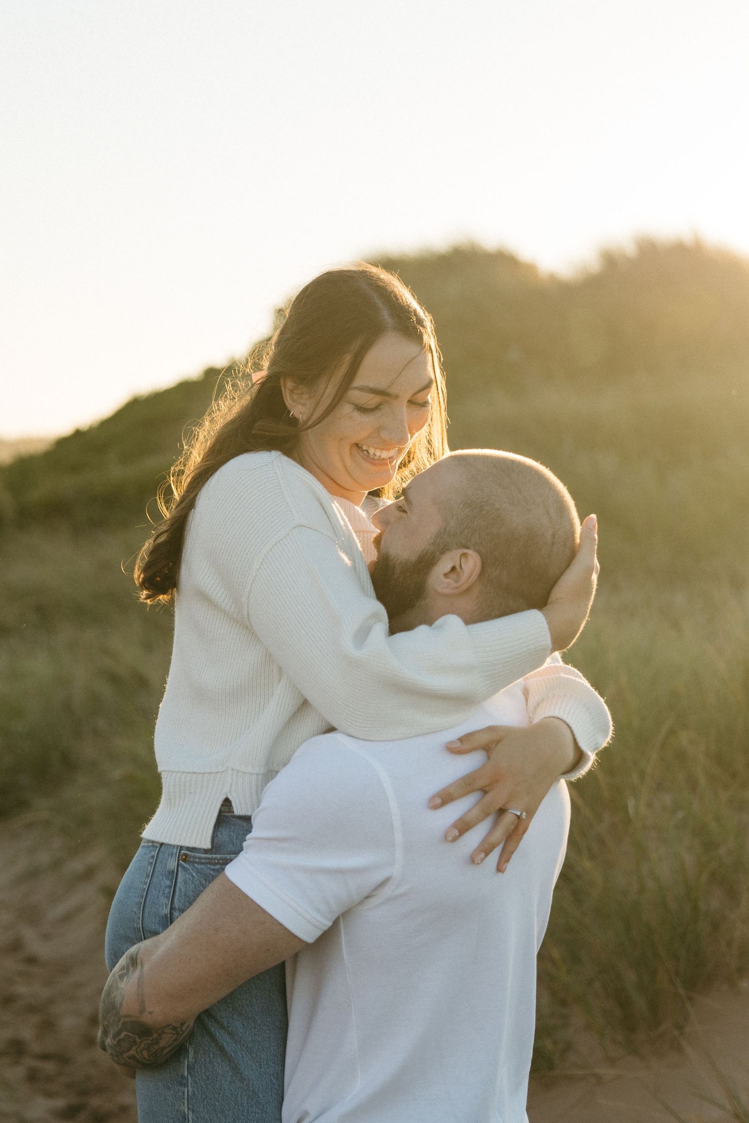 Romantic couple embraces in warm sunset light on sandy dunes with tall grass in background.